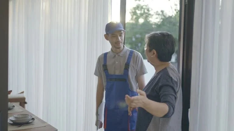 A young male worker in a blue uniform and cap talking to a woman by a large window in a restaurant or cafe setting.