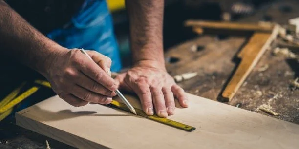 Person measuring a piece of wood with a tape measure in a woodworking shop.