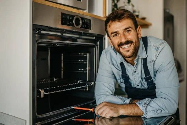 Man smiling in front of an open oven, holding a screwdriver, in a kitchen setting.