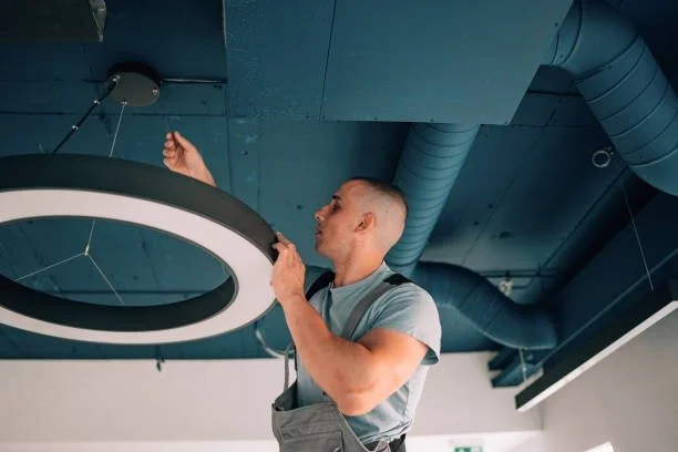 A man installing or repairing a circular ceiling light fixture in a modern space with exposed ceiling ducts.