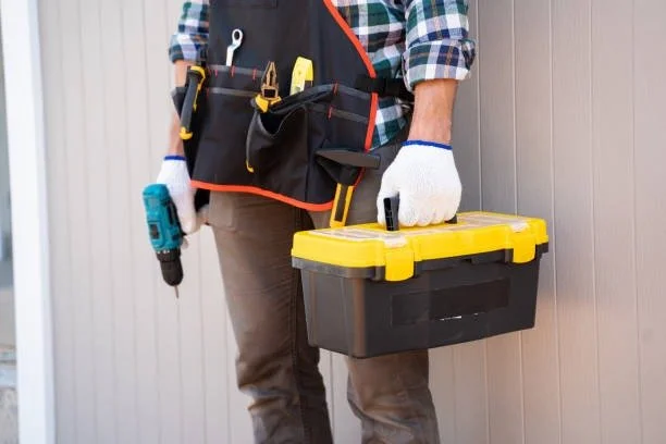 Person wearing work gloves holding a black and yellow toolbox with a yellow lid, standing next to a light-colored wooden wall, with a black tool belt containing various tools around their waist and a drill in their right hand.