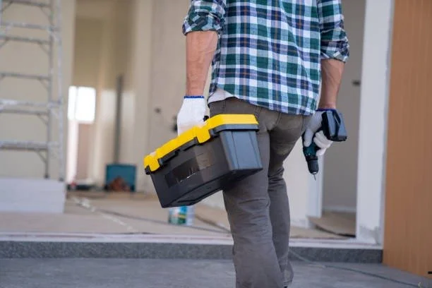 Person carrying a toolbox and holding a cordless drill inside a construction or renovation site.