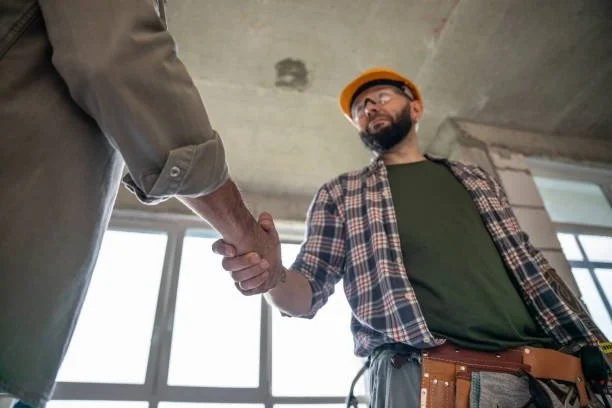 Construction worker with a beard wearing a yellow hard hat and plaid shirt shaking hands with another person