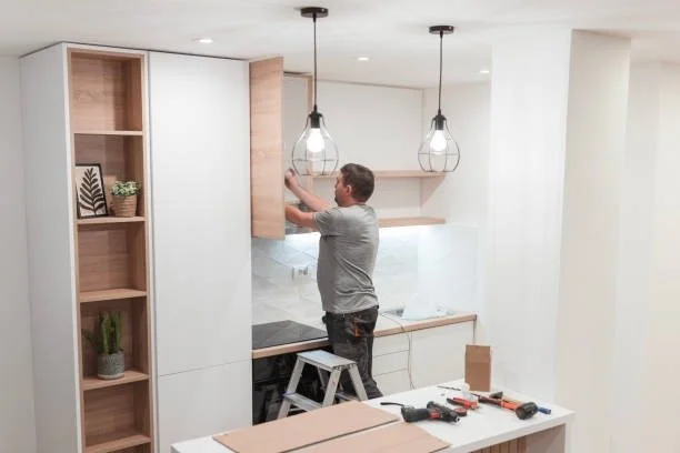 Person installing kitchen cabinets in a modern kitchen with pendant lights and a marble backsplash.
