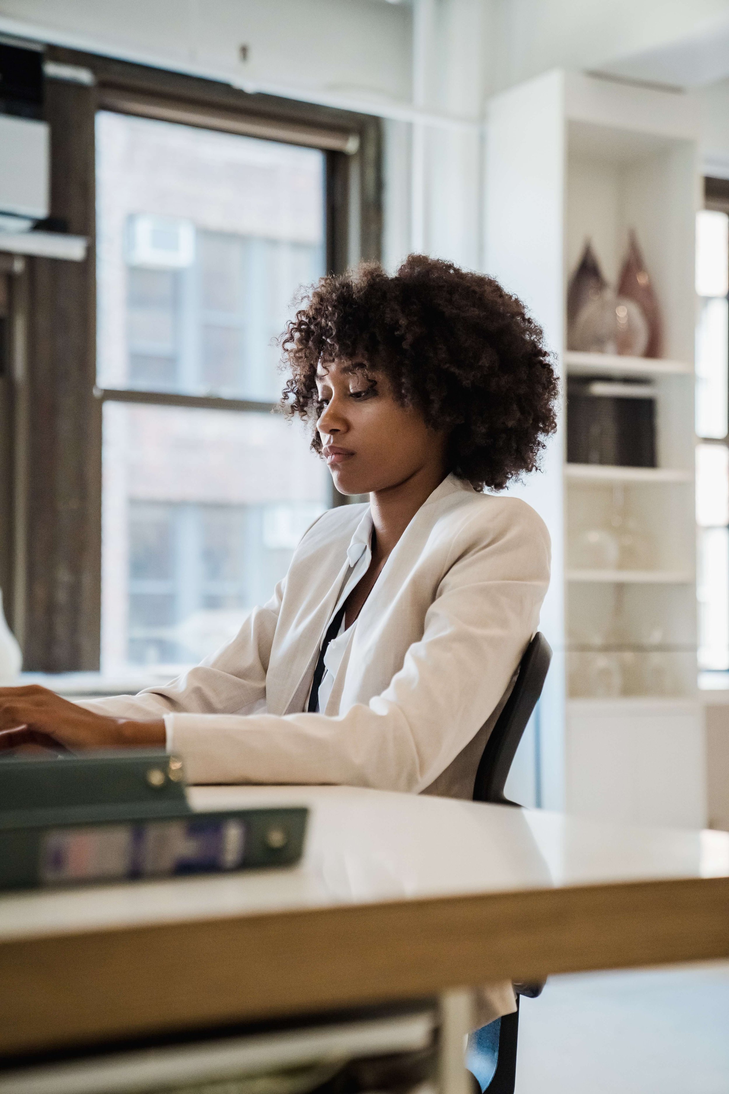 A woman with curly hair, wearing a beige blazer, sitting at a desk in a modern office, working on a laptop.