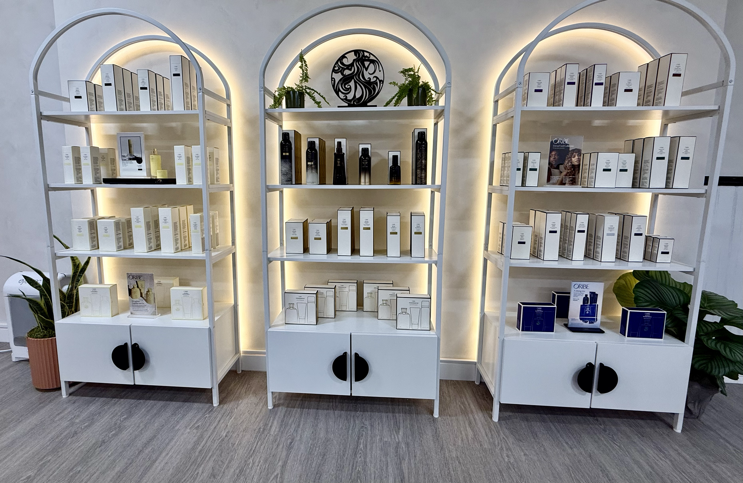 A white display shelf with various skincare or cosmetic products, illuminated with soft lighting, flanked by potted plants, set against a white wall.