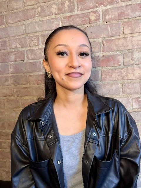 A woman with long dark hair, wearing makeup, earrings, a gray shirt, and a black leather jacket, standing in front of a brick wall.