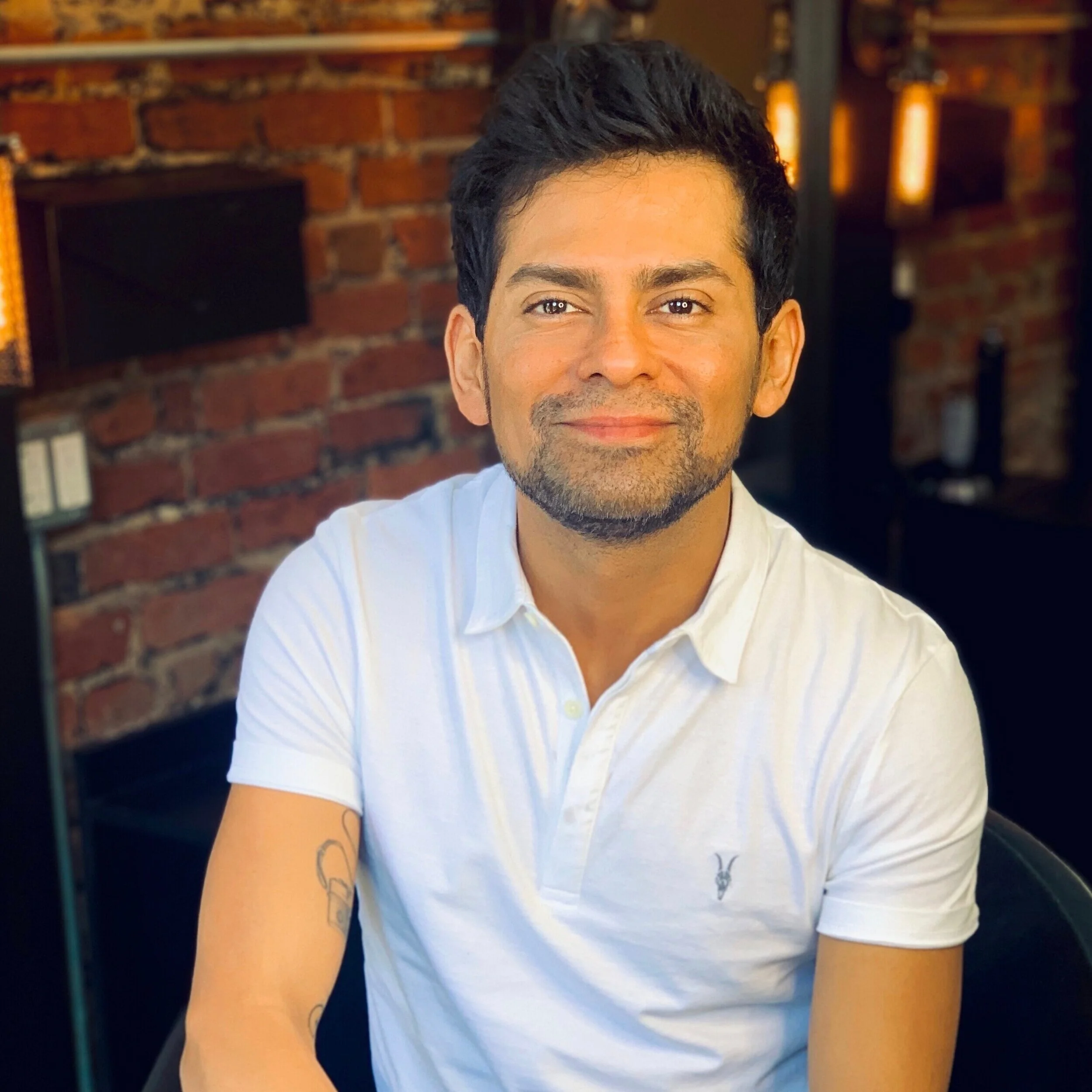Close-up of a smiling man with dark hair, wearing a white shirt, with a brick wall and warm lighting in the background.