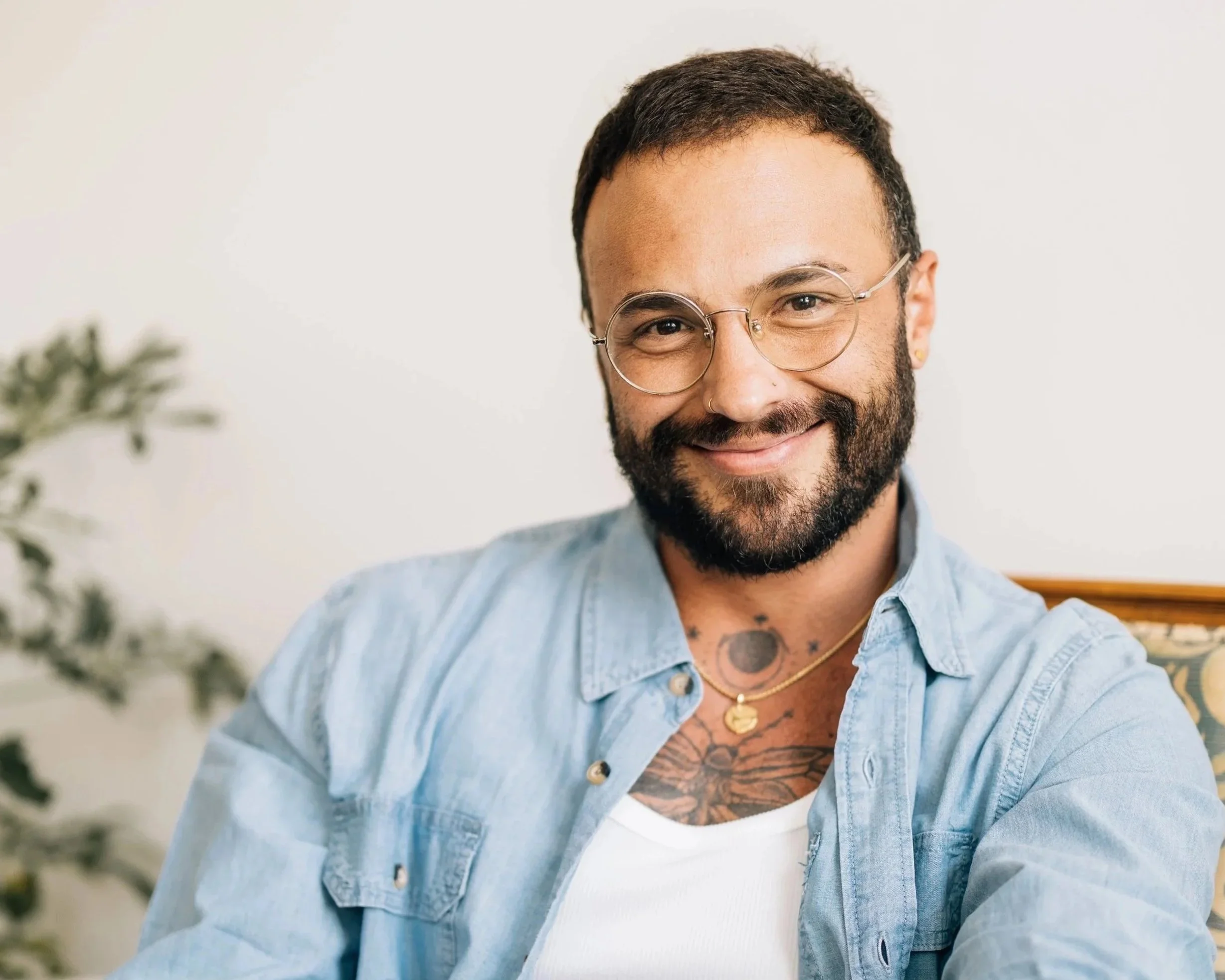 A smiling man with glasses, a beard, and tattoos on his chest, wearing a light blue denim shirt over a white T-shirt, sitting indoors near a Christmas tree.