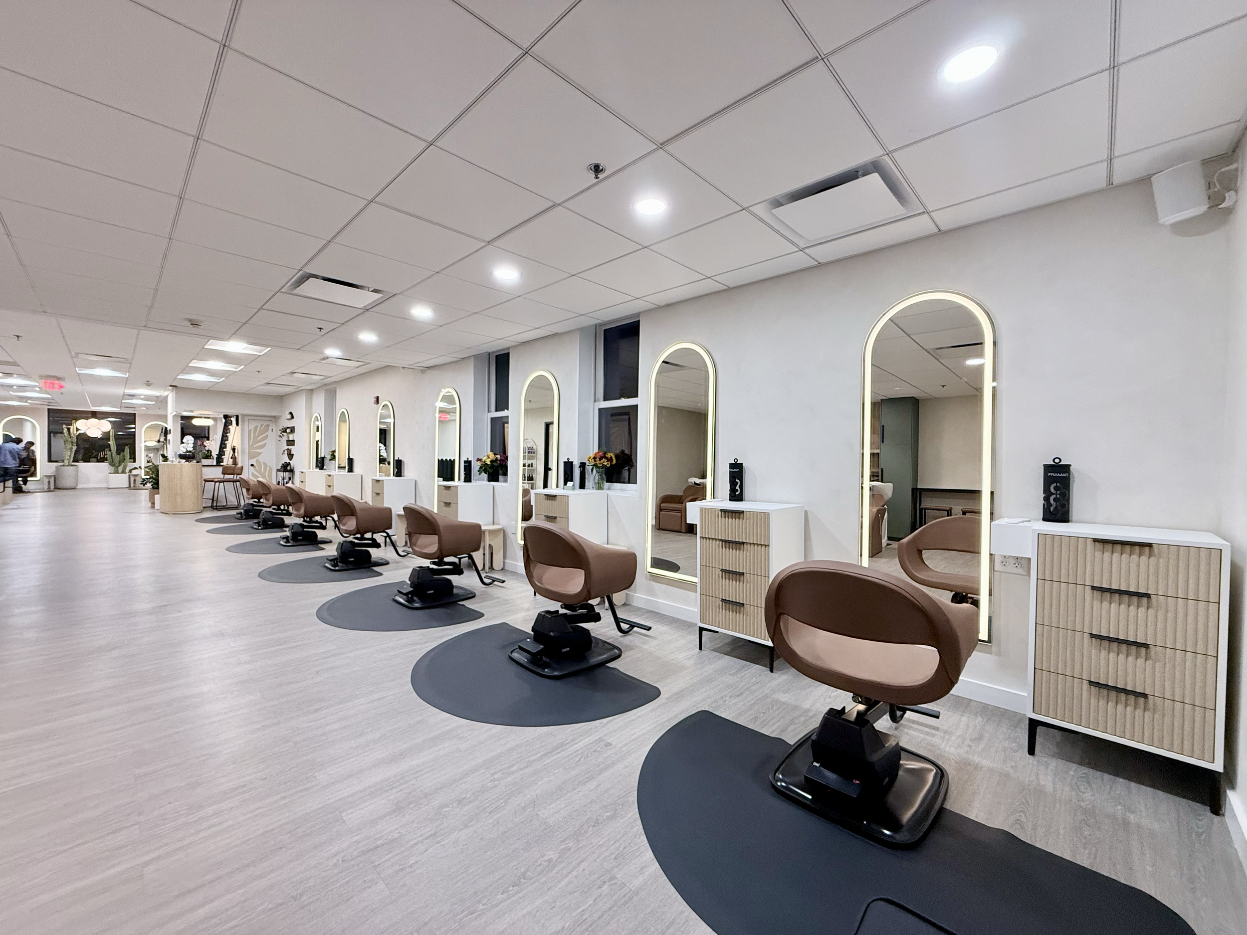 A modern hair salon interior with multiple styling stations. Each station has a brown salon chair, a mirror with illuminated borders, and a small cabinet. The salon has a light-colored floor and white walls, with decorative plants and flowers on some