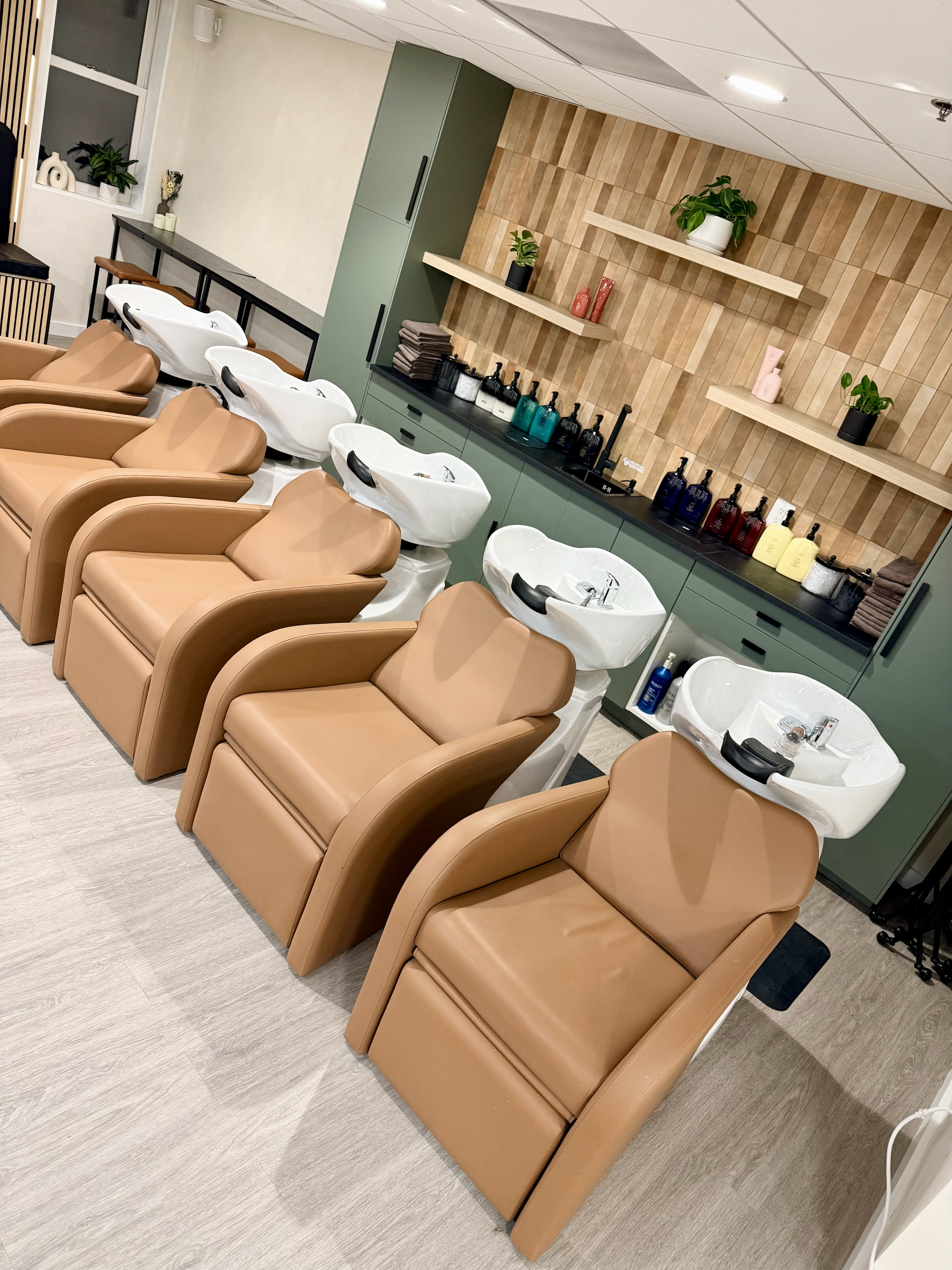 Hair washing station with four brown reclining chairs, each with a white sink in front. Behind the chairs, there are light green cabinets, shelves with potted plants, and decorative items.