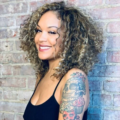 Woman with curly hair and a colorful tattoo on her upper arm, smiling against a brick wall background.