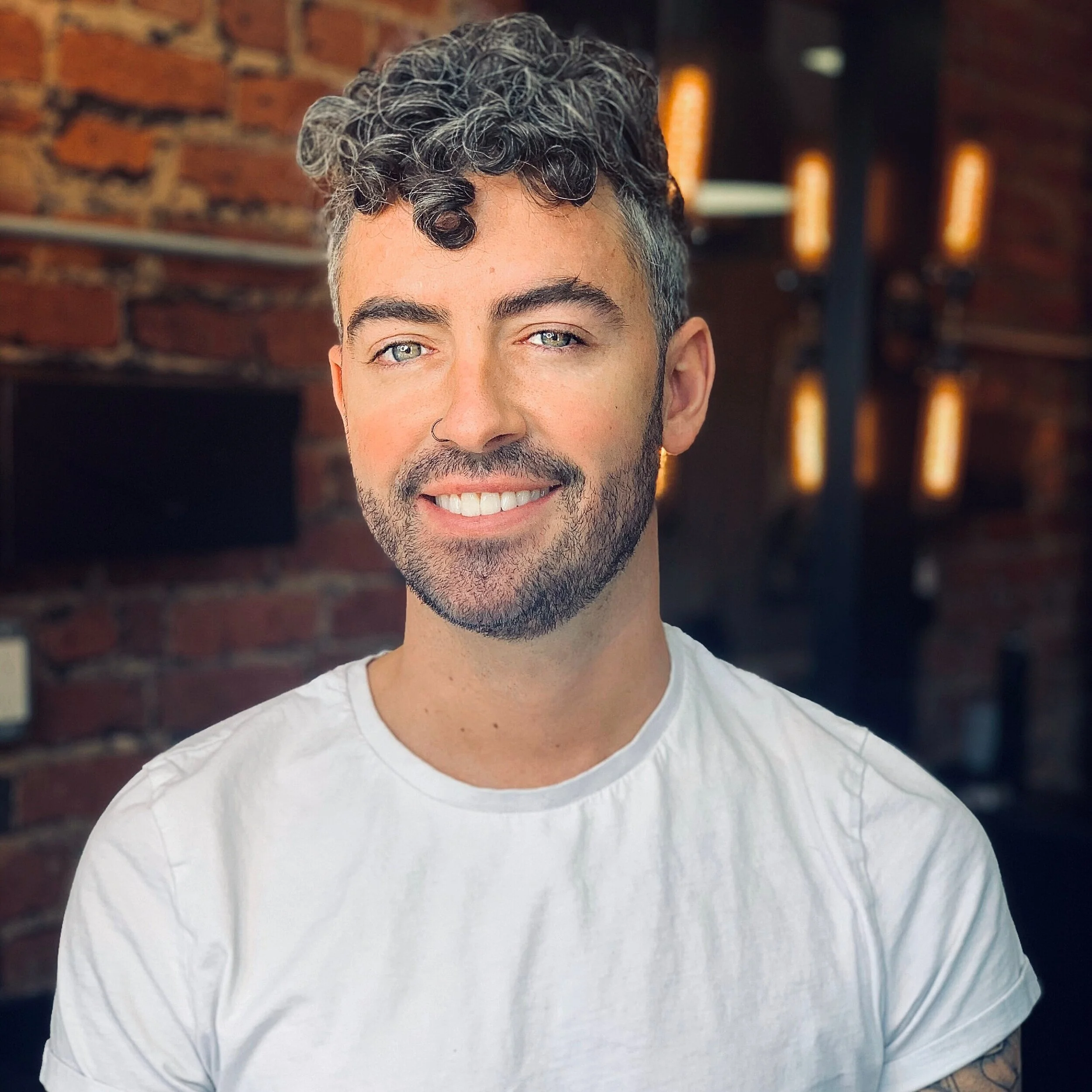 Close-up of a man with curly, gray-highlighted hair and a beard, wearing a white T-shirt in a dimly lit indoor space with exposed brick wall and warm lighting.