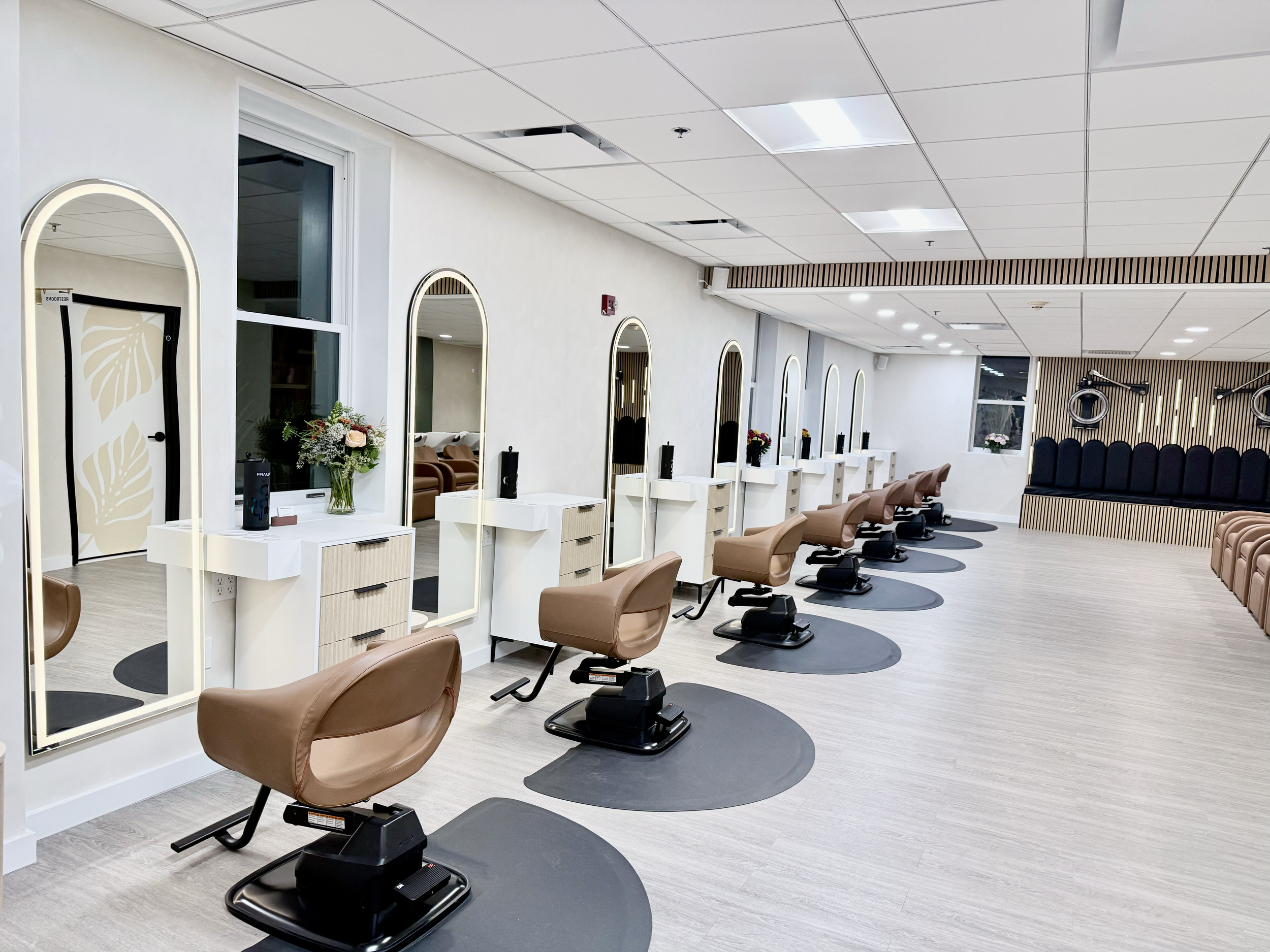 Modern hair salon with styling stations, large mirrors, and salon chairs arranged in a row. The space features a clean, neutral color palette and decorative elements such as flowers and striped wall panels.