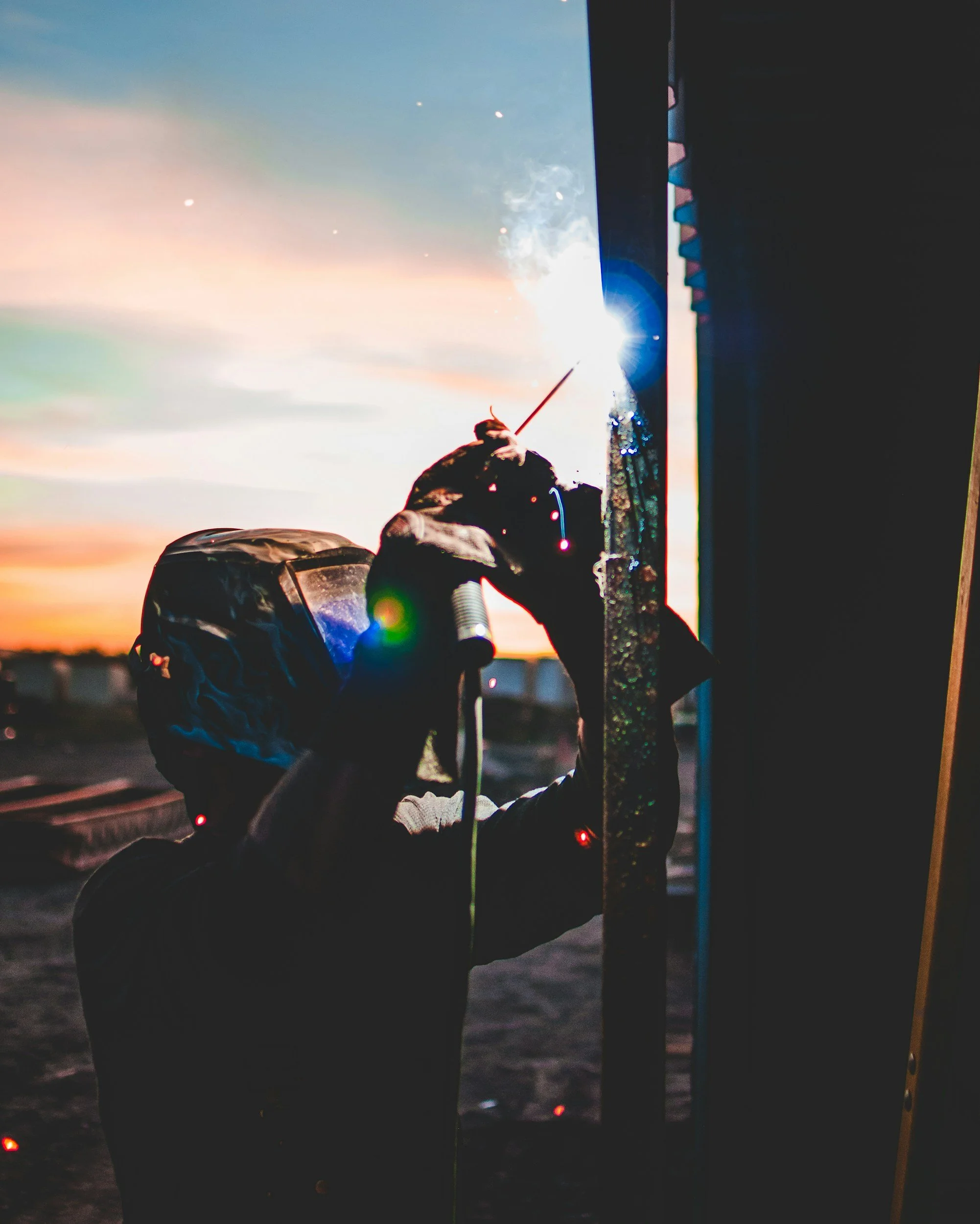 A worker welding metal outdoors during sunset with sparks and bright light.
