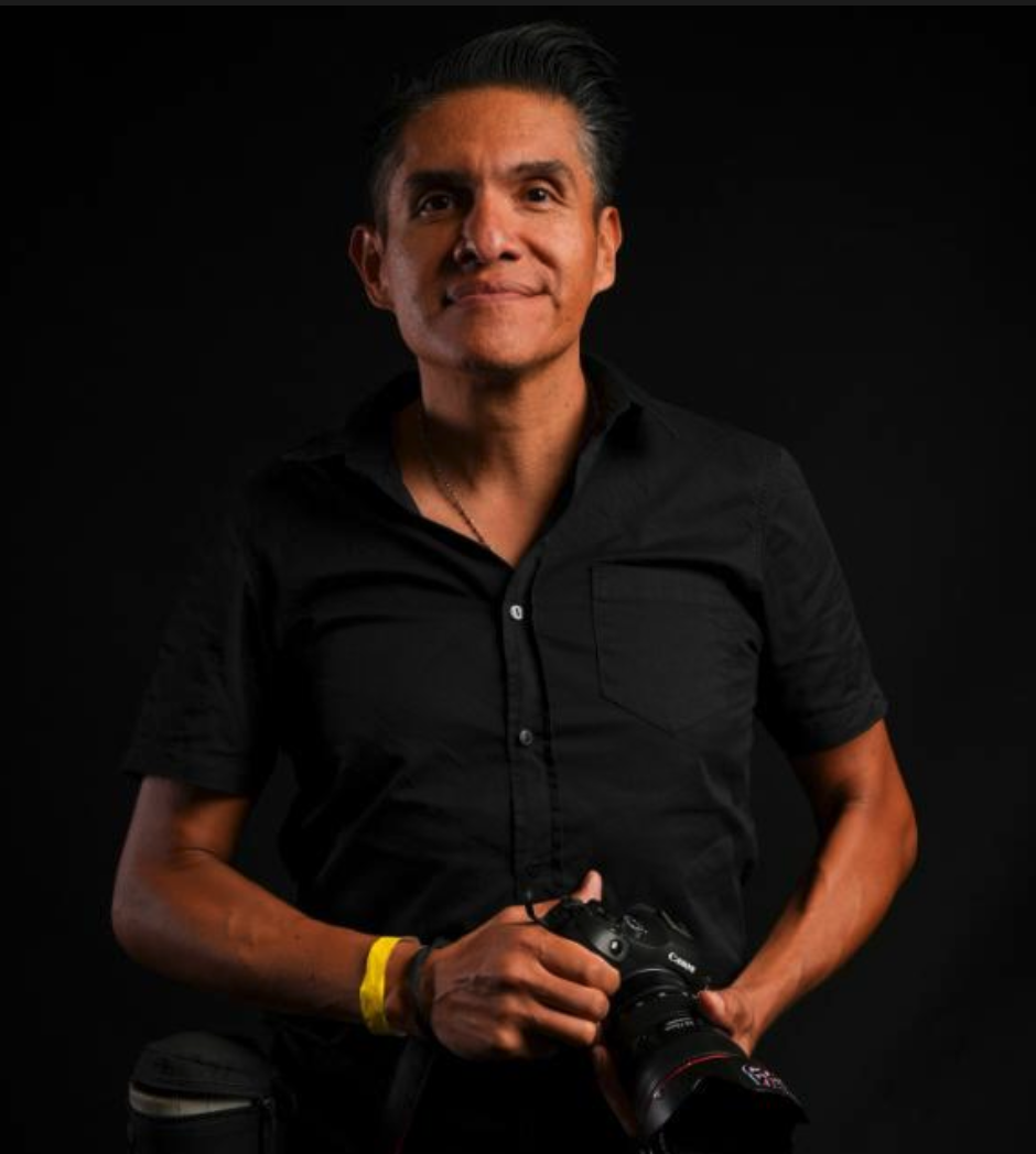Un hombre joven con cabello oscuro, ropa negra y sonriendo, sostiene una cámara profesional con fondo negro.