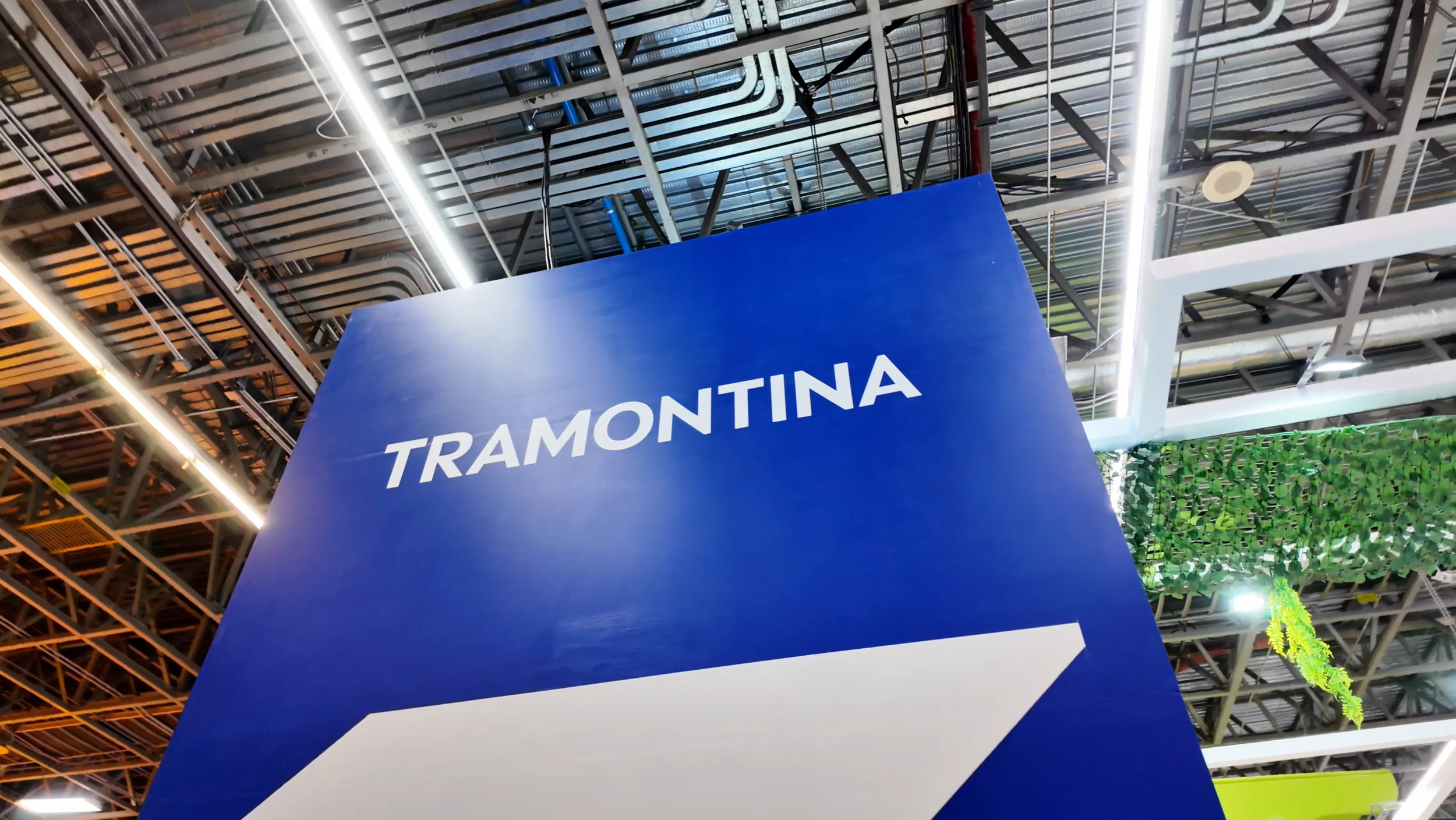 Se observa un cartel azul con la palabra 'TRAMONTINA' en letras blancas, colocado en un espacio interior con techo de estructura metálica y luces LED.