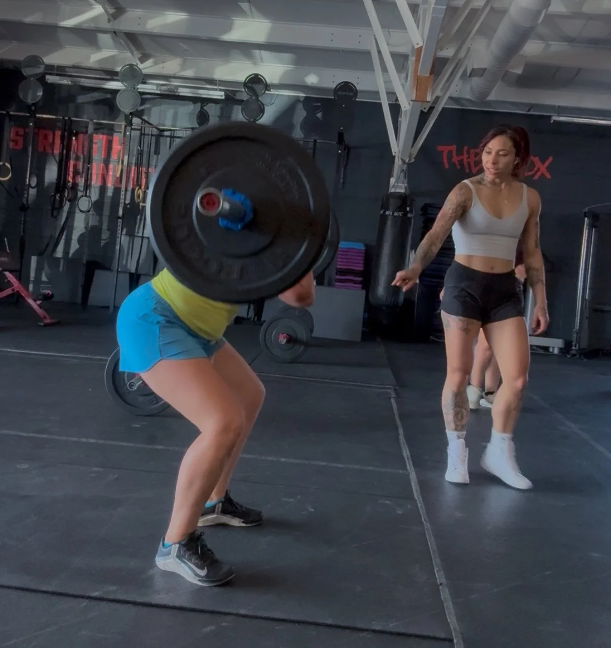A woman performing a barbell squat in a gym, with a trainer observing nearby.
