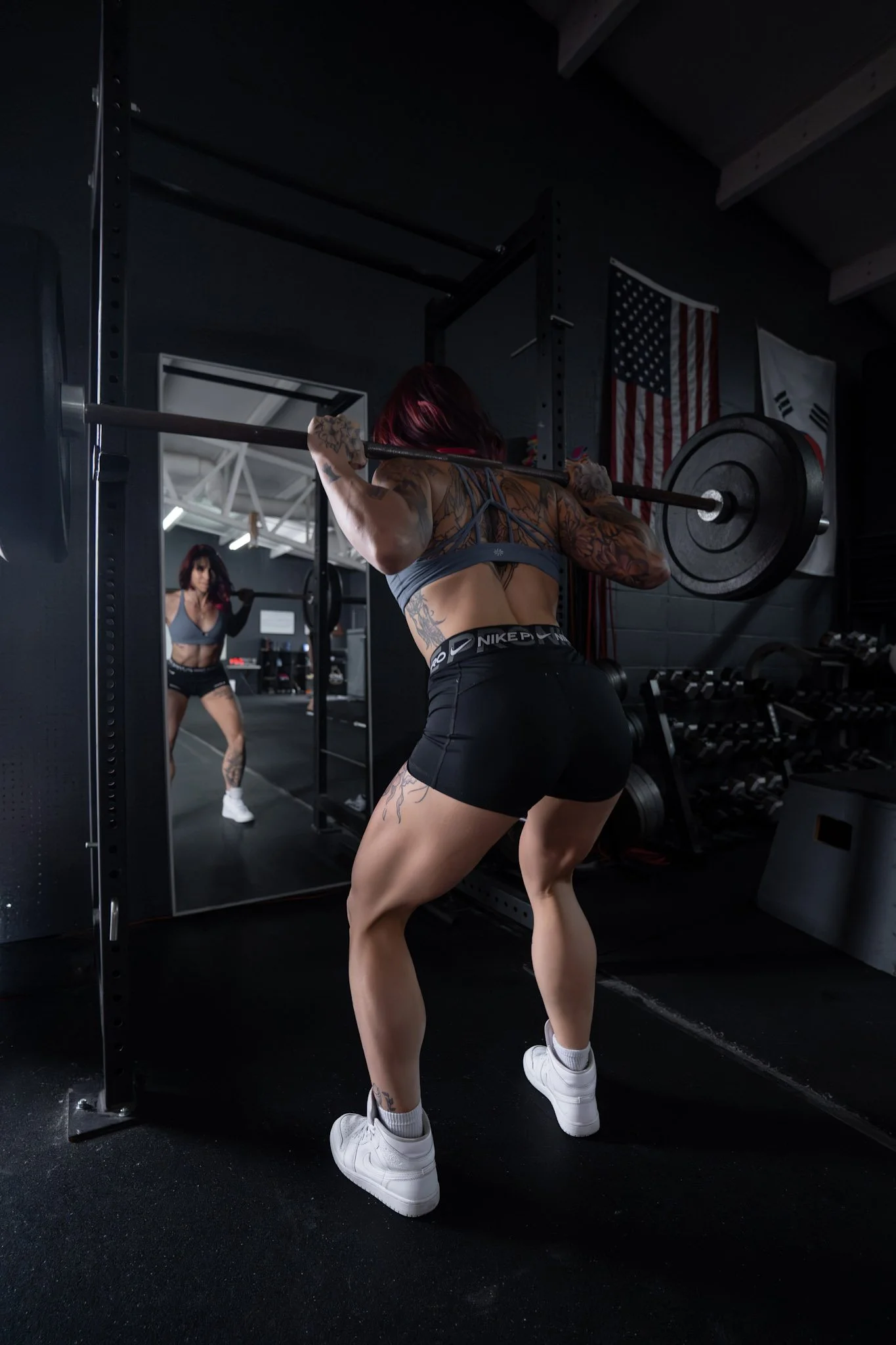 A woman with tattoos working out with a barbell in a gym, her reflection seen in a mirror.
