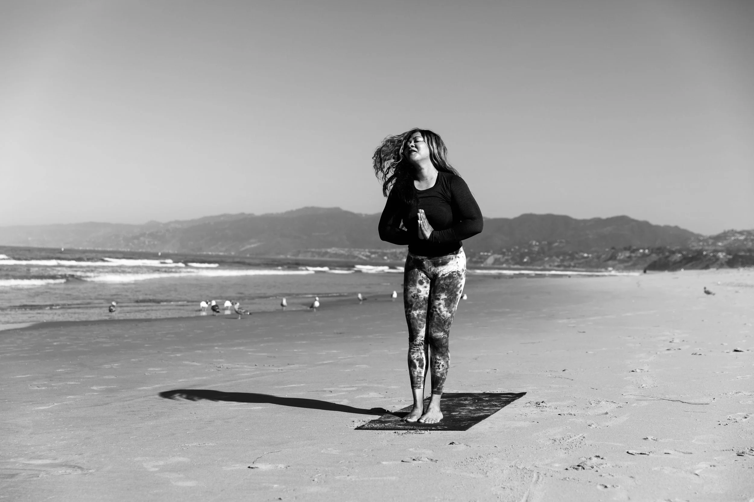 A woman practicing yoga on a beach, standing on a mat with her palms together in prayer position, ocean waves and mountains in the background.