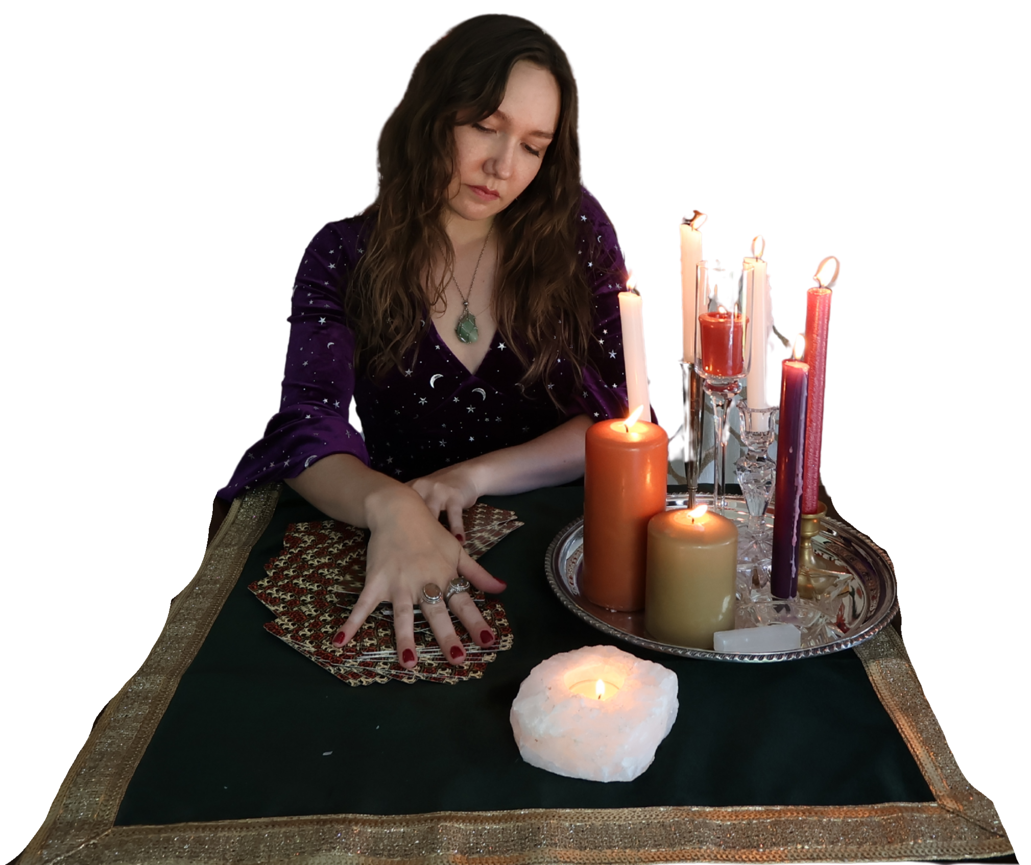 A woman with long brown hair, wearing a purple dress with moon and star patterns, sits at a table with tarot cards and candles. The table has a dark cloth with gold trim, and a silver tray holds various lit candles of different colors and sizes. A white candle holder with a lit candle and a clear crystal are also on the table.