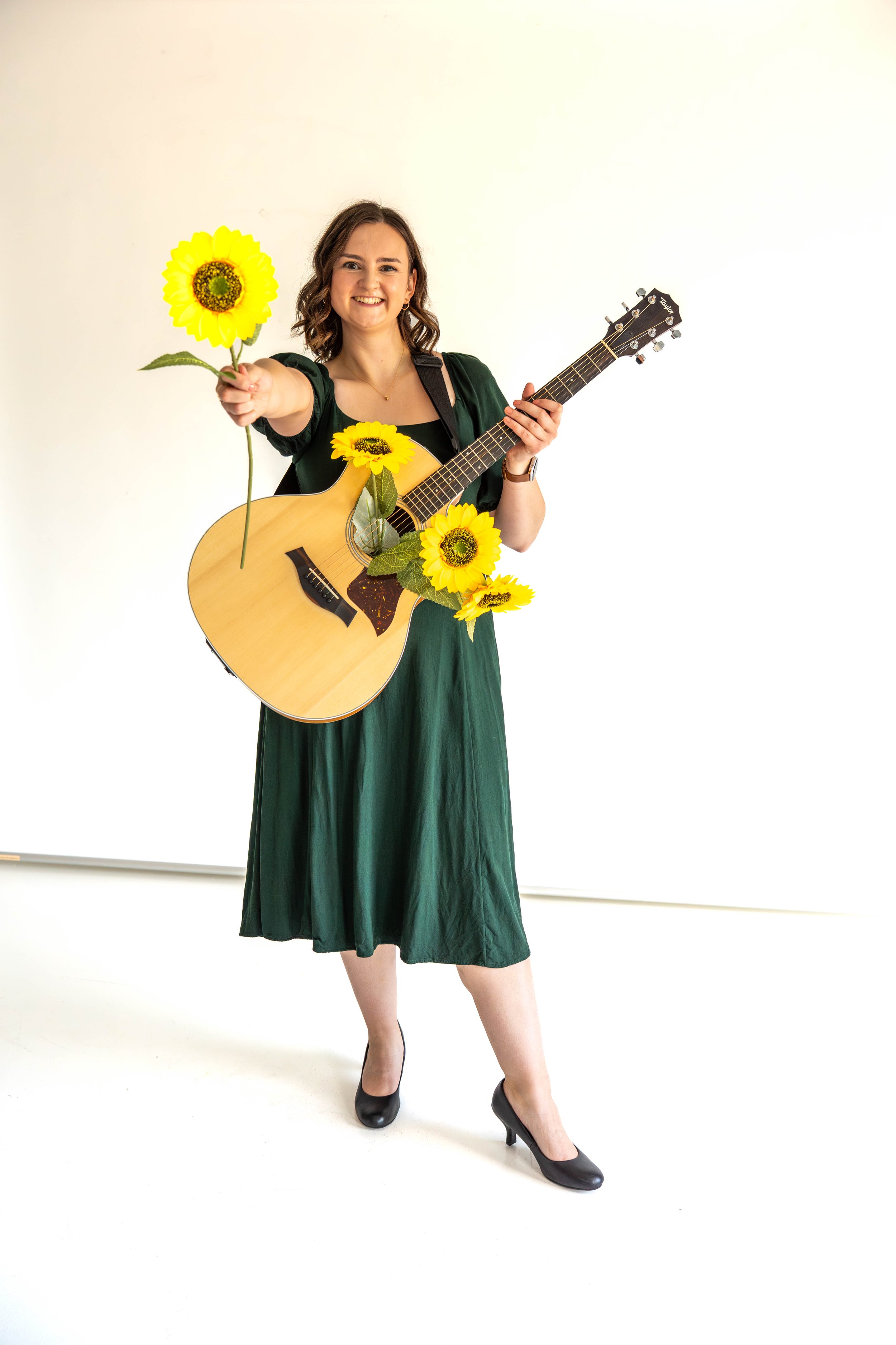Gabi Frens, a white woman with brown hair and brown eyes, stands with a guitar strapped to her. The guitar's sound hole holds three faux sunflowers, and Gabi smiles while holding out another sunflower.