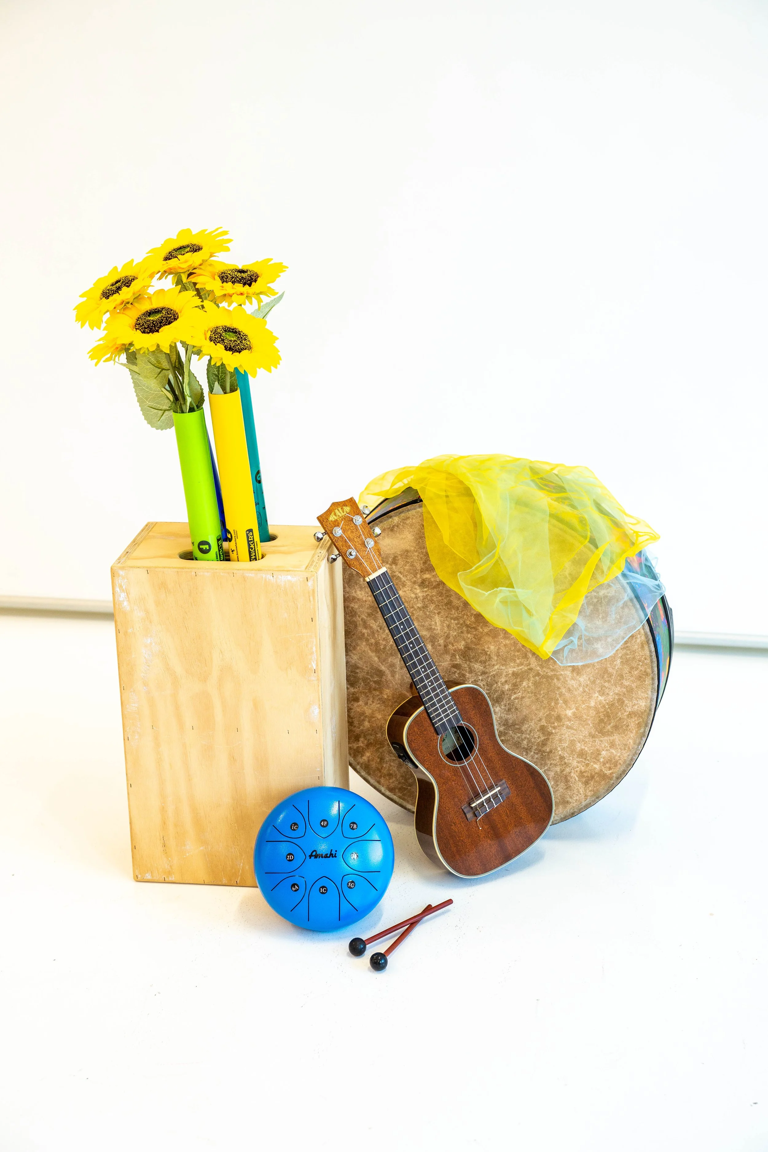Image of musical instruments including a tongue drum, a ukulele, a floor drum propped on it's side with scarves on top - all leaned against a wooden box holding sunflowers and Boomwhackers