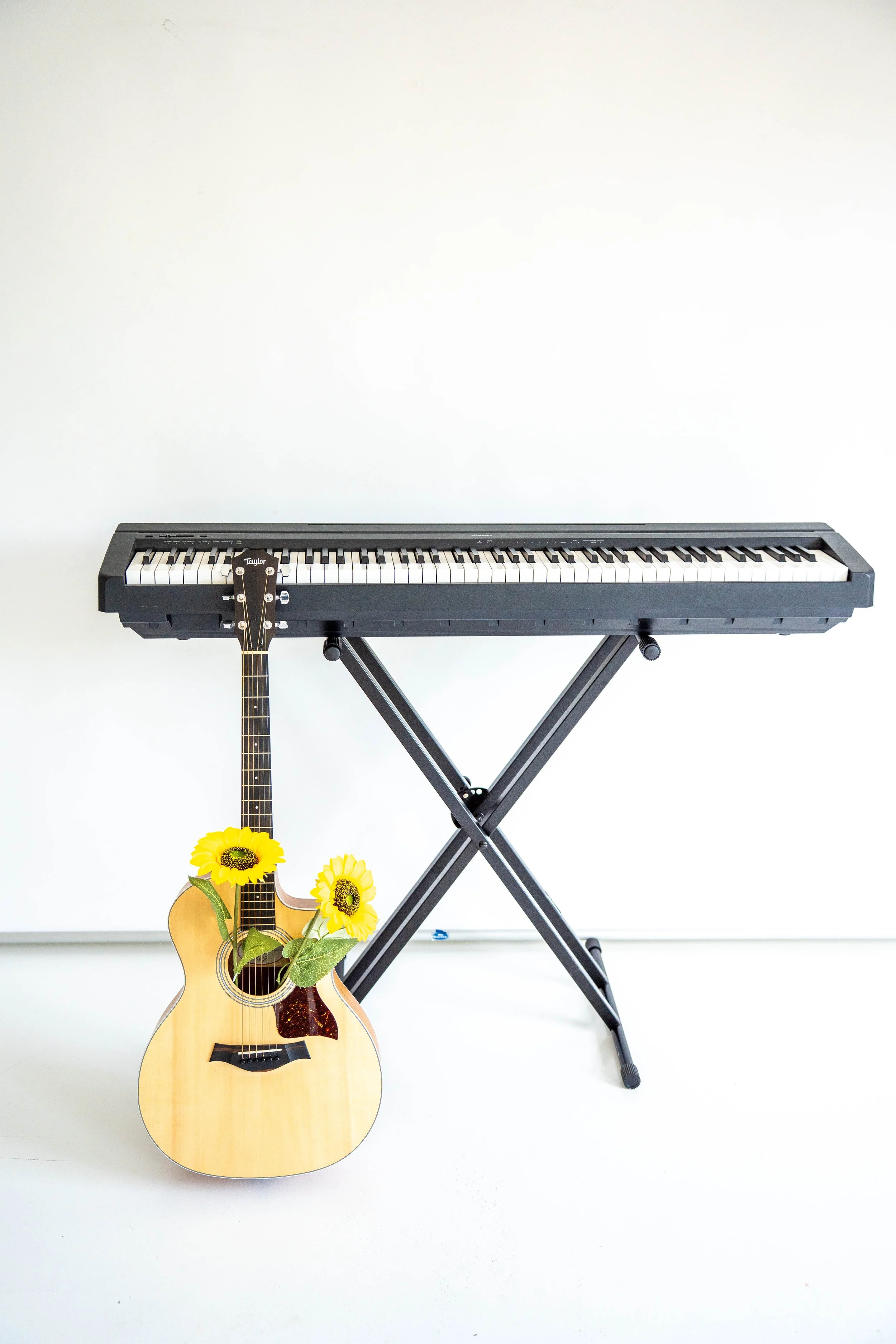 A keyboard set on a stand with a guitar leaning up against it. The guitar's sound hole has two faux sunflowers in it.