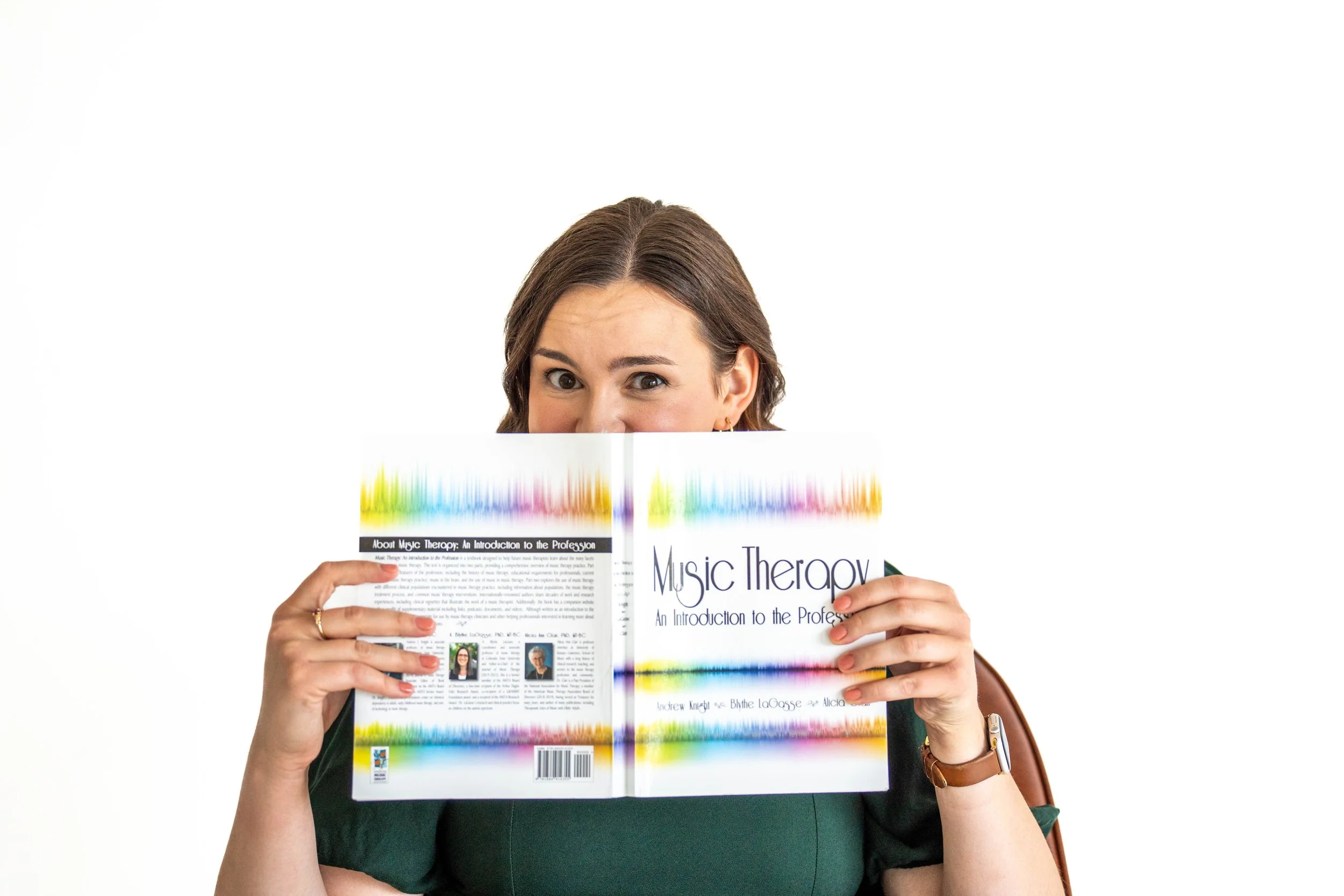 Gabi Frens, a white woman with brown hair, sits holding a copy of "Music Therapy: An Introduction to the Profession" to partially cover her face