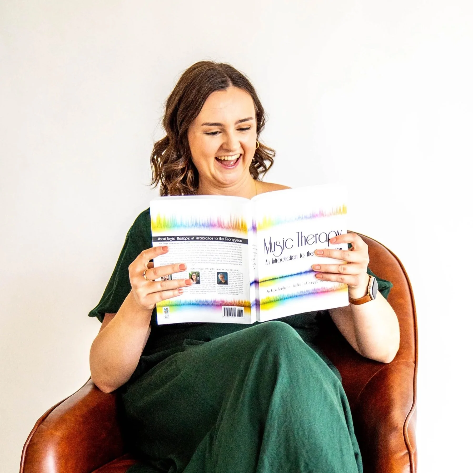 Gabi Frens, a white woman with brown hair and brown eyes, sits in a chair reading "Music Therapy: An Introduction to the Profession." She is smiling.