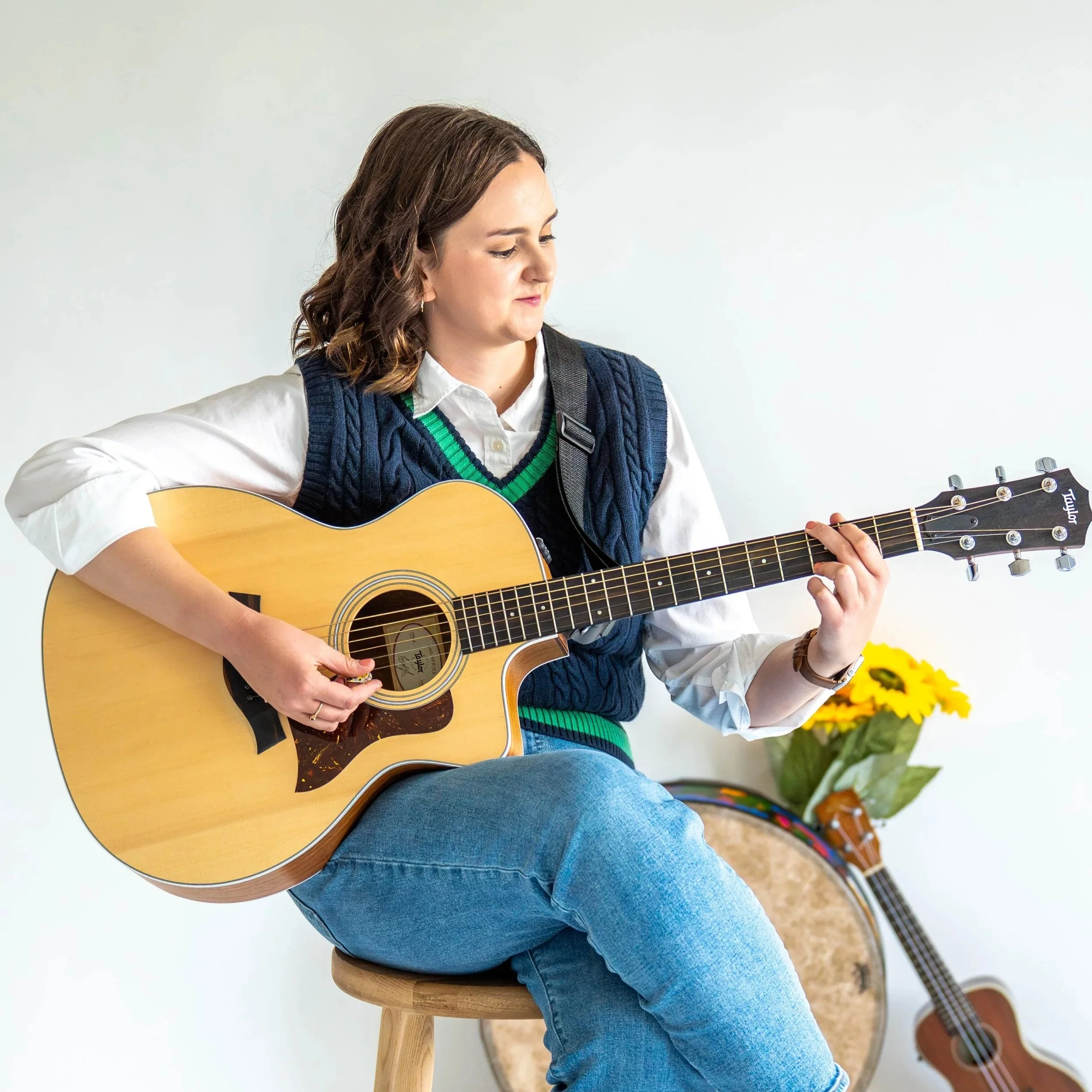 Gabi Frens, a white woman with brown hair and brown eyes, sits on a wooden stool holding a guitar and looking at it while playing. The background features a floor drum, a ukulele, and some fake sunflowers leaned against the wall.