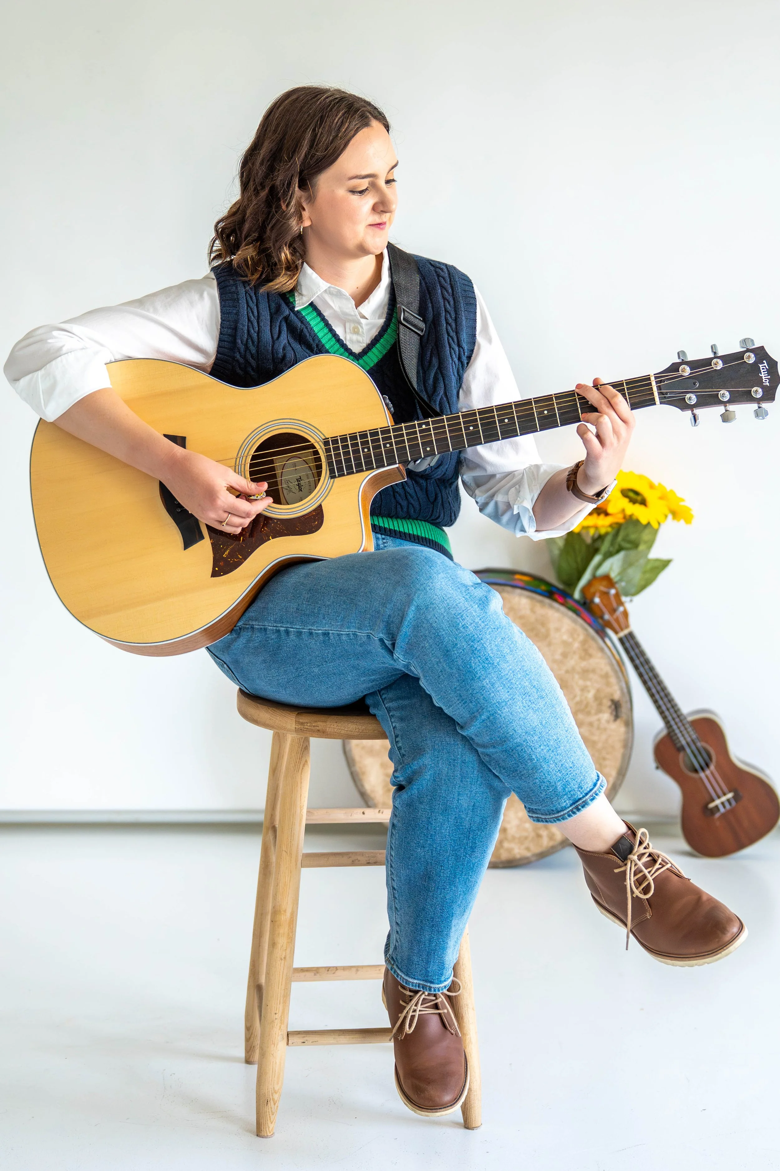 Gabi Frens, a white woman with brown hair and brown eyes, sits on a wooden stool holding a guitar and looking at it while playing. The background features a floor drum, a ukulele, and some fake sunflowers leaned against the wall.