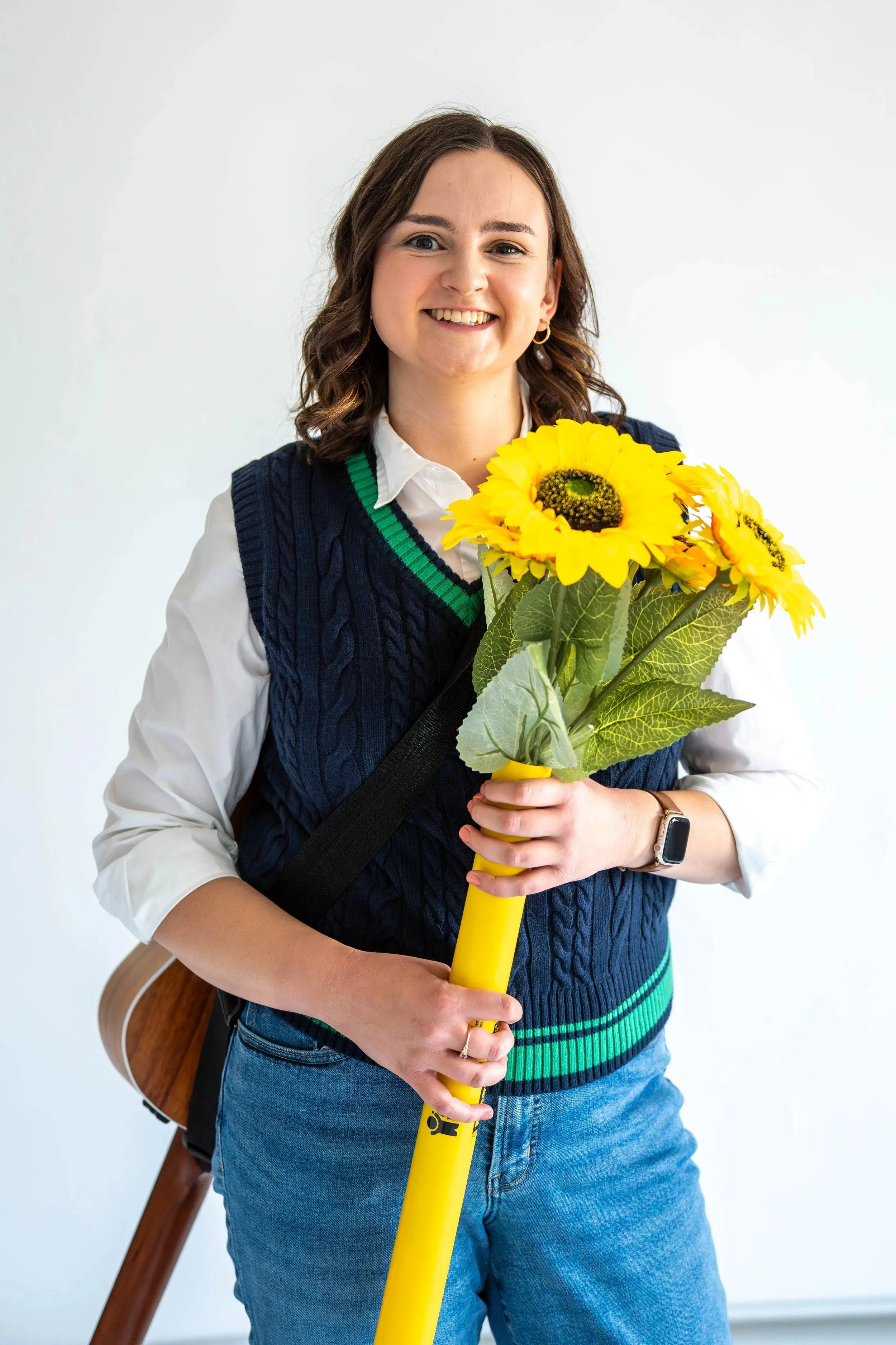 Gabi Frens, a white woman with brown hair and brown eyes stands holding a bouquet of fake sunflowers inside a Boomwhacker instrument. She has a guitar on her back
