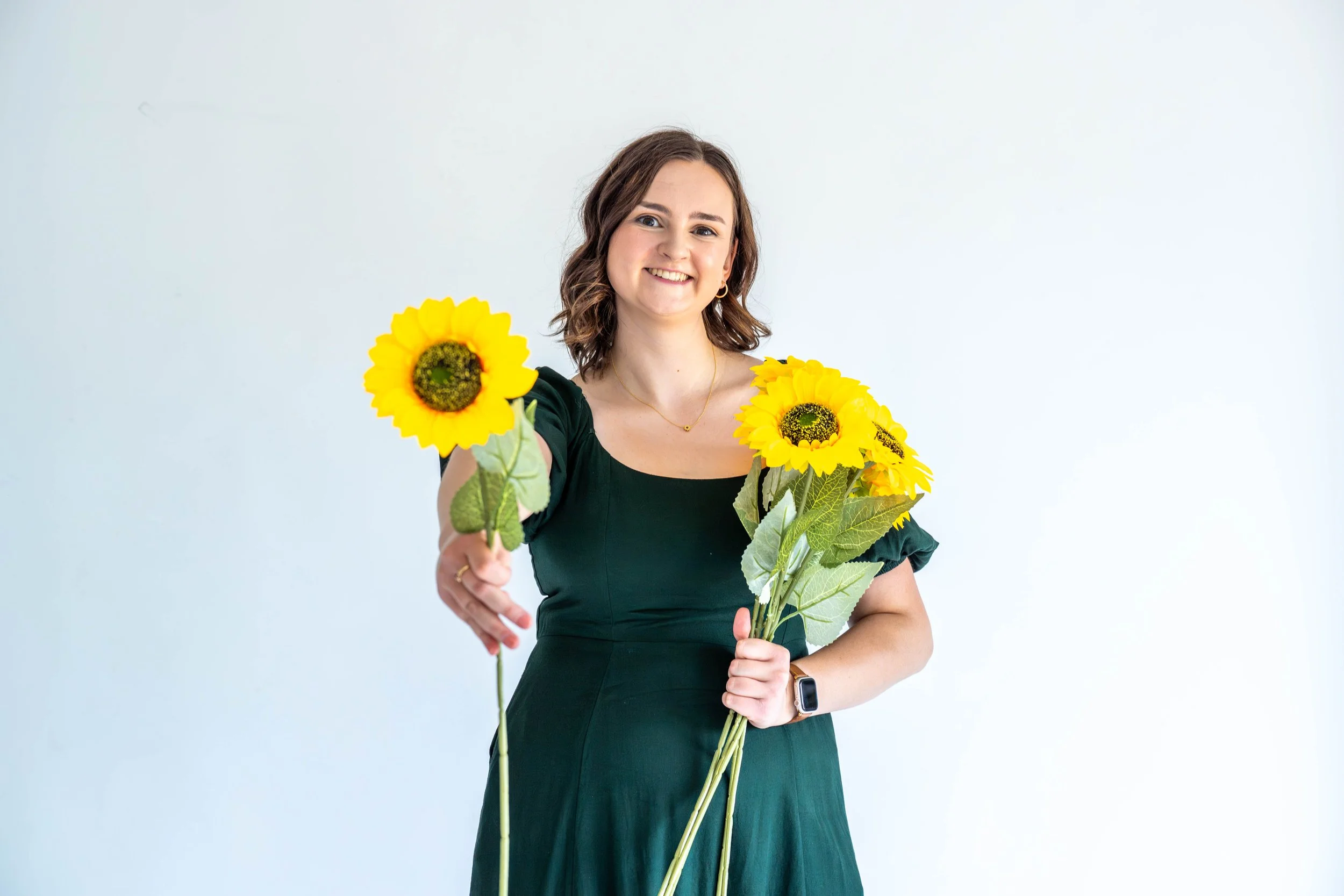Gabi Frens, a white woman with brown hair and brown eyes smiles while holding out a fake sunflower