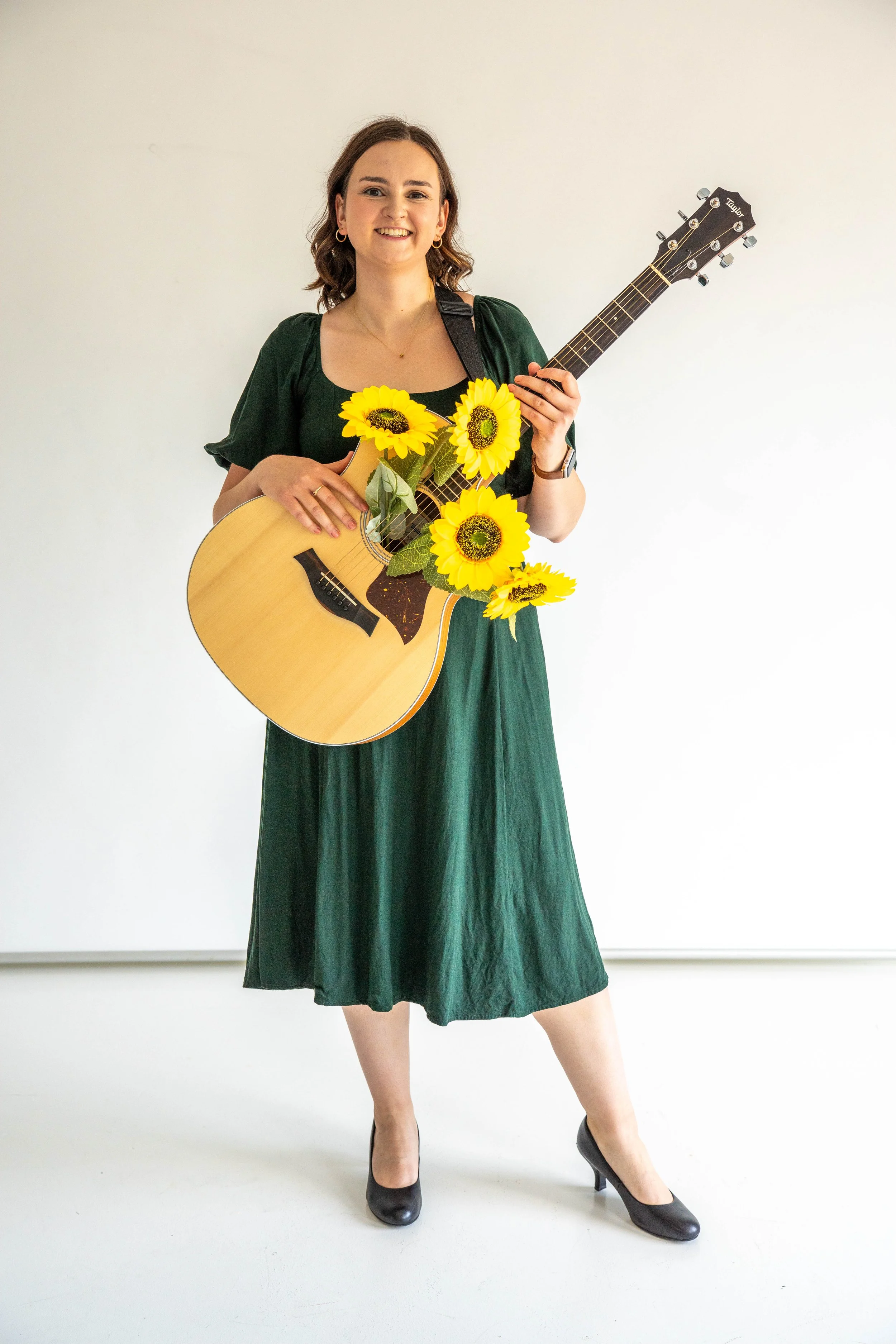 Gabi Frens, a white woman with brown hair and brown eyes, stands smiling. She holds a guitar that is strapped across her body. The guitar has four faux sunflowers coming out of the sound hole.