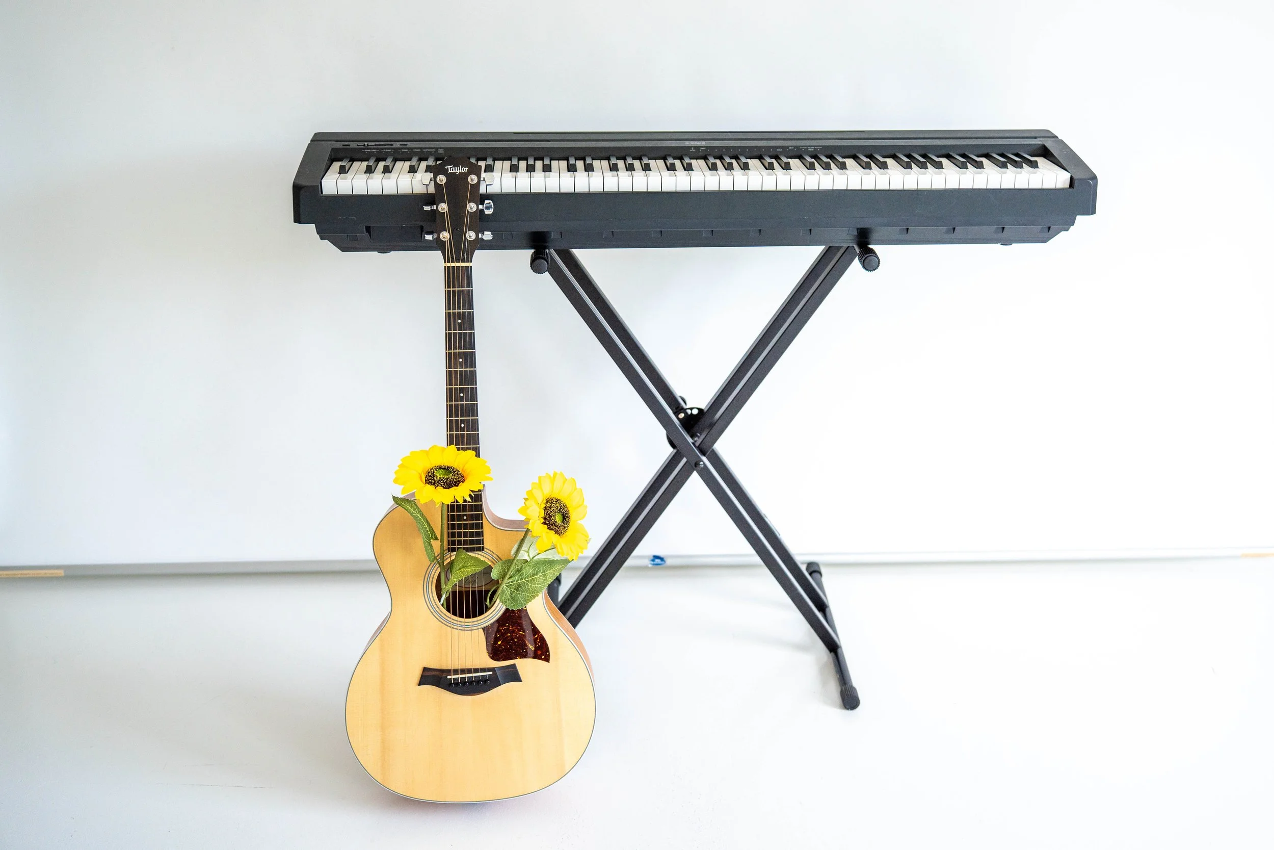 A keyboard set on a stand with a guitar leaning up against it. The guitar's sound hole has two faux sunflowers in it.