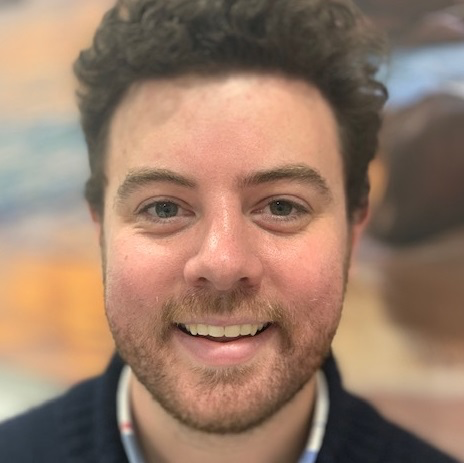Close-up of a smiling young man with curly brown hair and a beard, wearing a dark sweater