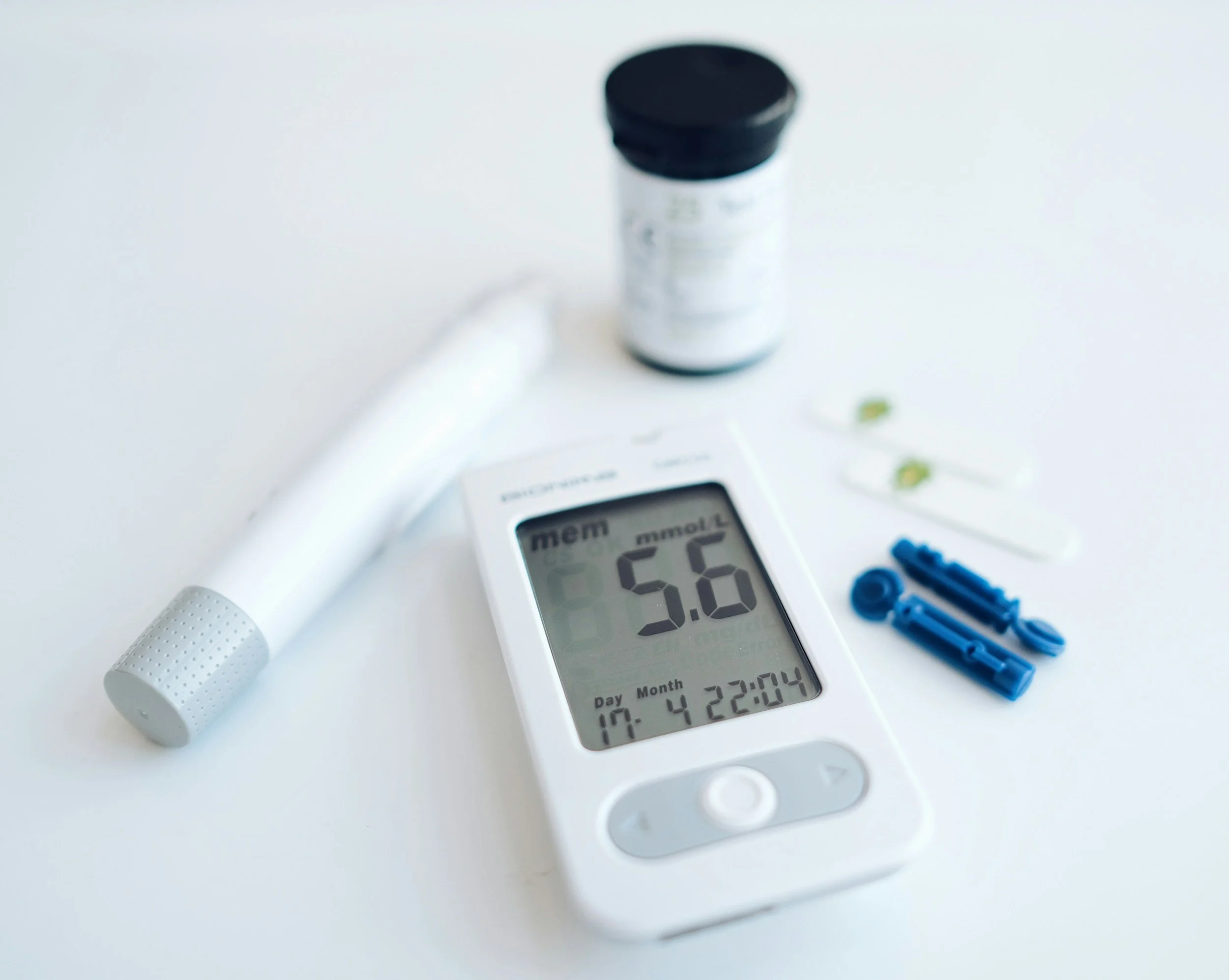 A digital glucose meter displaying a reading of 5.6 mmol/L, placed on a white surface along with a white blood glucose control solution bottle, a lancing device, and test strips.