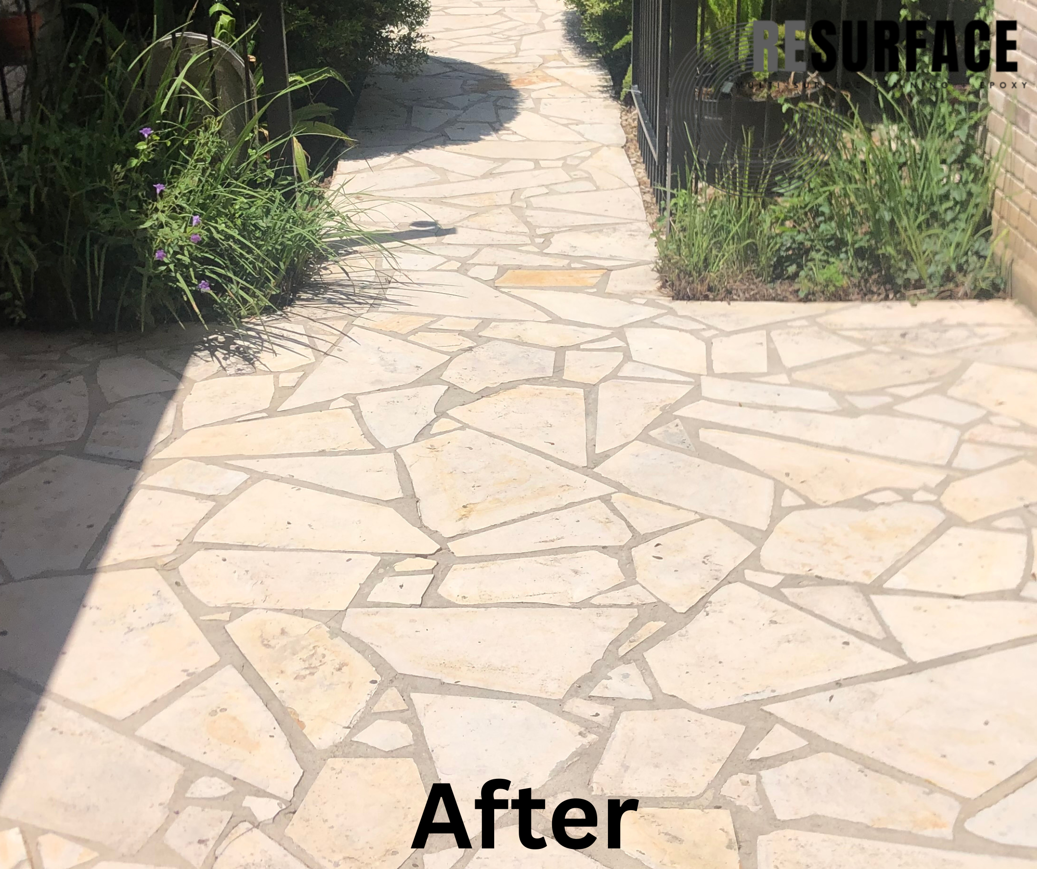Photo of a newly installed stone patio with irregular-shaped paving stones, bordered by plants and a black metal fence, with sunlight casting shadows.