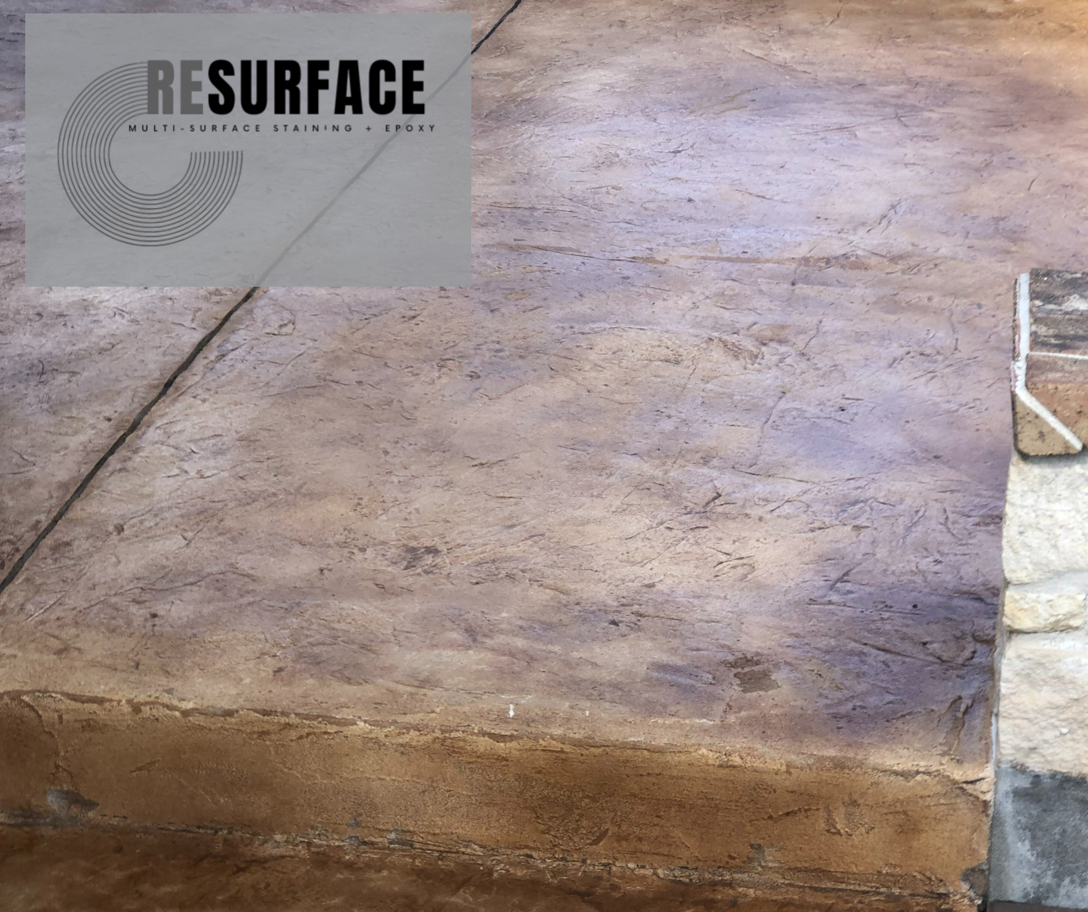 Close-up view of a textured stone or tile surface with a corner of a stone wall visible on the right, and a logo watermark reading RESURFACE in the top left corner.
