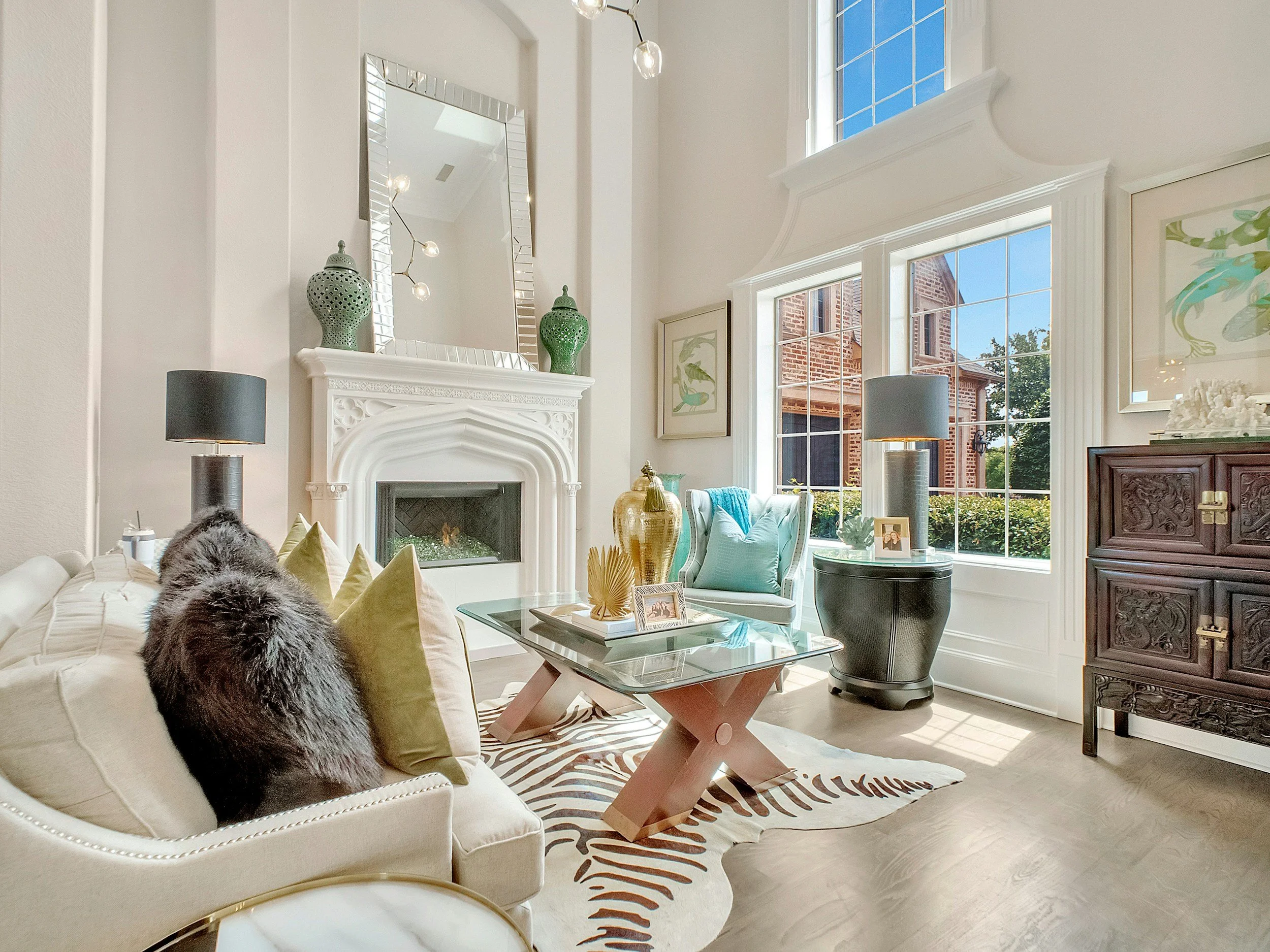 Bright living room with white walls, large windows, a fireplace with a mirror above, and a mix of modern and traditional furniture. A cream sofa with pillows and fur, glass coffee table, and decorative vases.