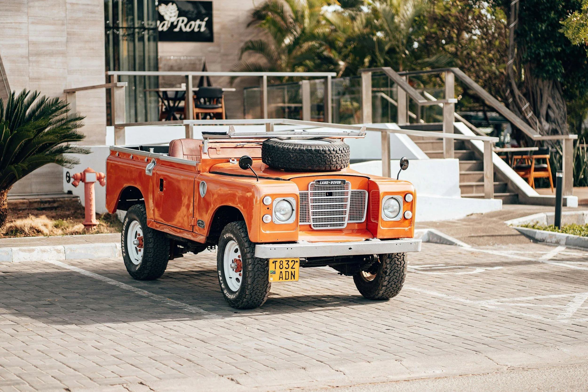 An orange vintage Land Rover parked in a lot in front of a building with outdoor seating and plants.