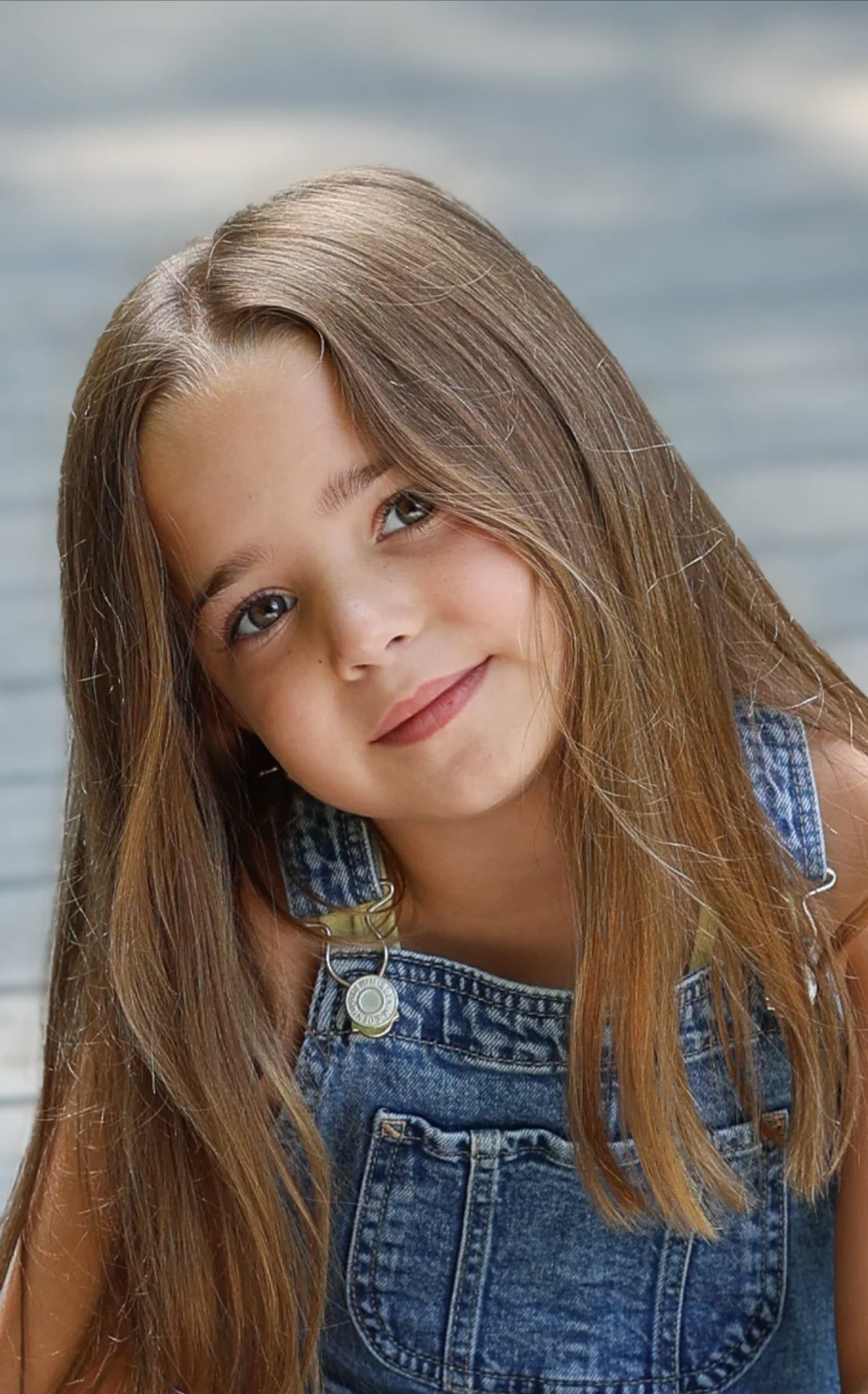 Close-up of a young girl with long, light brown hair, wearing denim overalls, smiling softly against a wooden background.