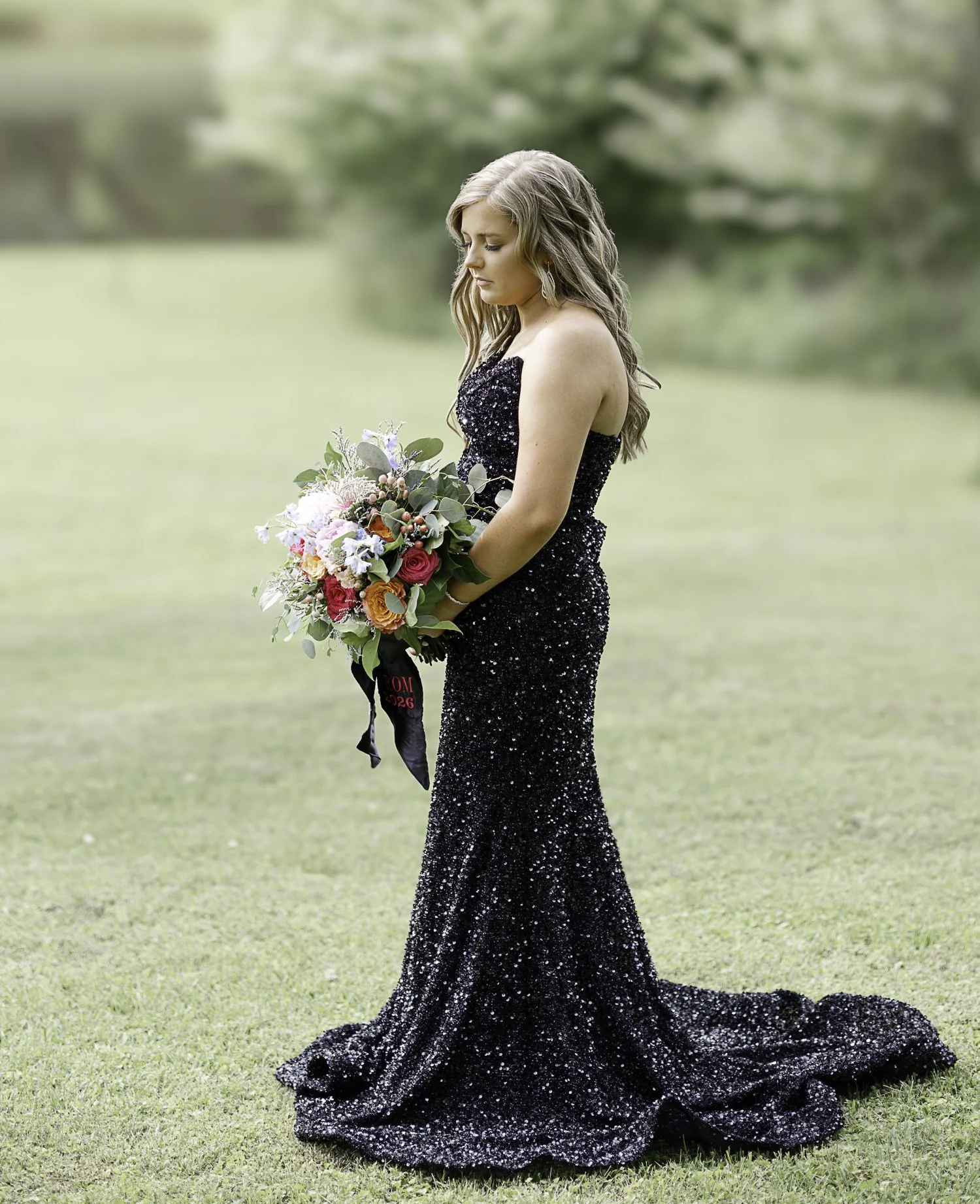 A woman in a black, sparkling, floor-length gown holding a bouquet of colorful flowers in a grassy outdoor setting.