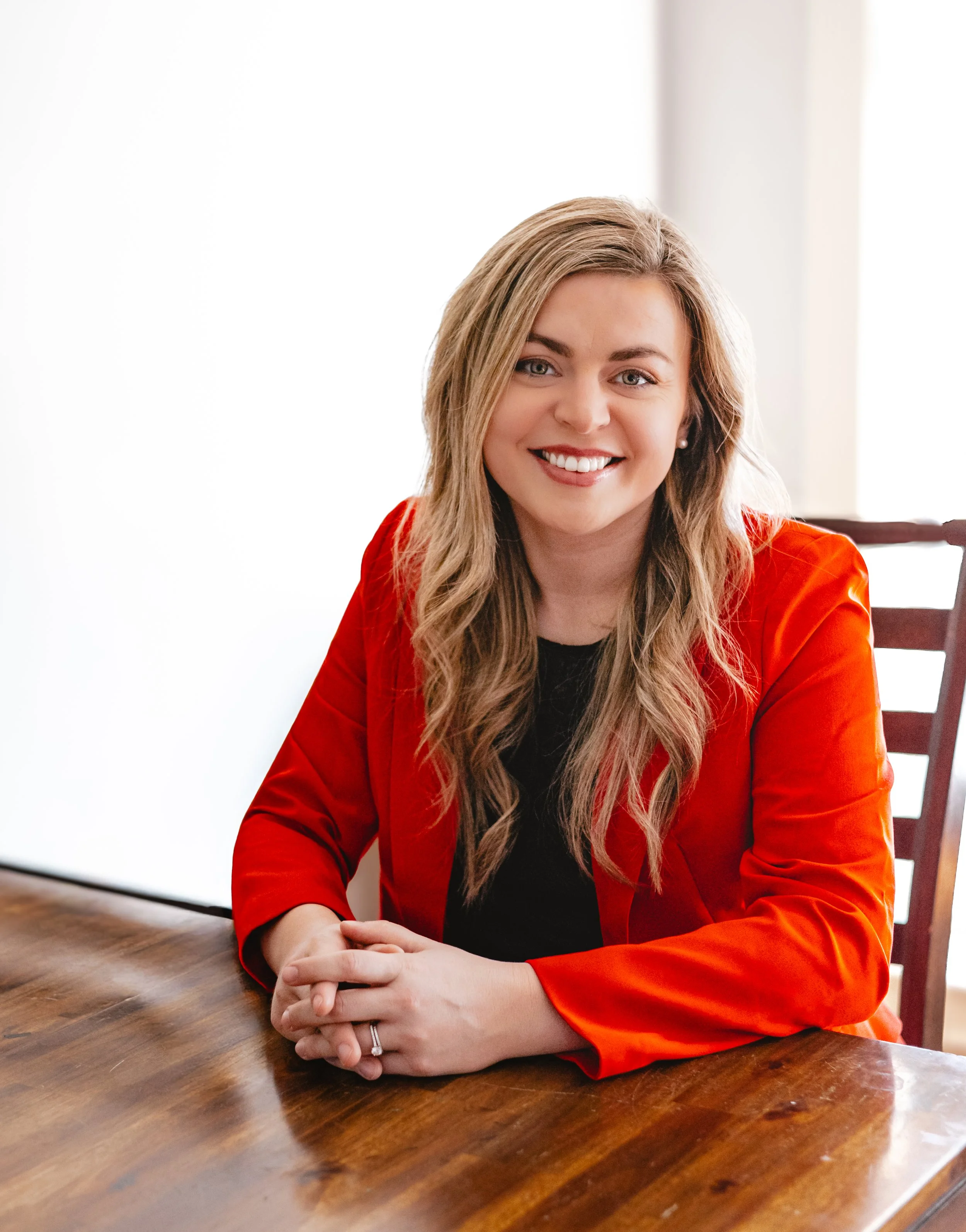 A woman with long blonde hair, wearing a red jacket, sitting at a wooden table, smiling at the camera.