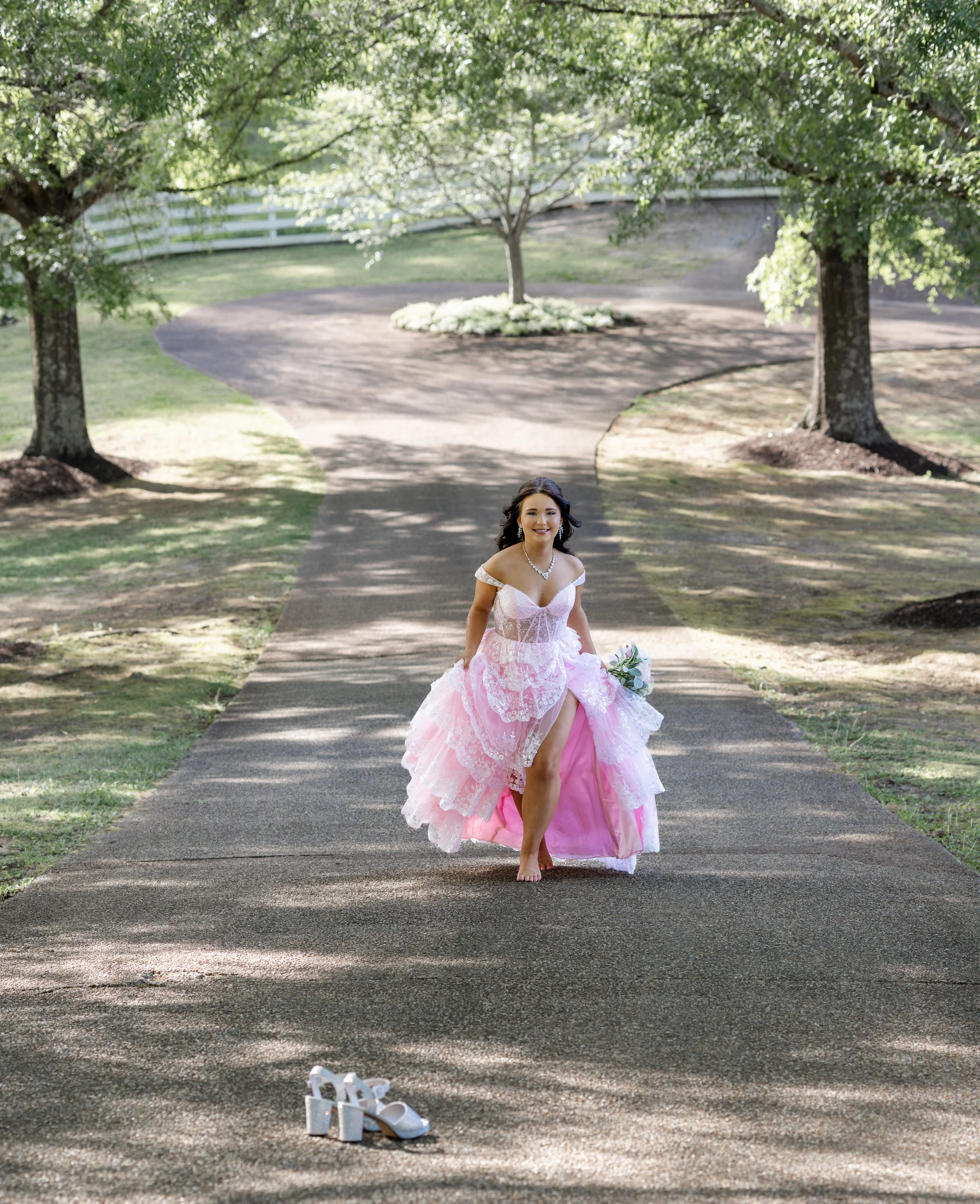 A woman in a pink lace dress standing barefoot on an outdoor pathway surrounded by trees, with white high heels and a small bouquet of flowers on the ground in front of her.