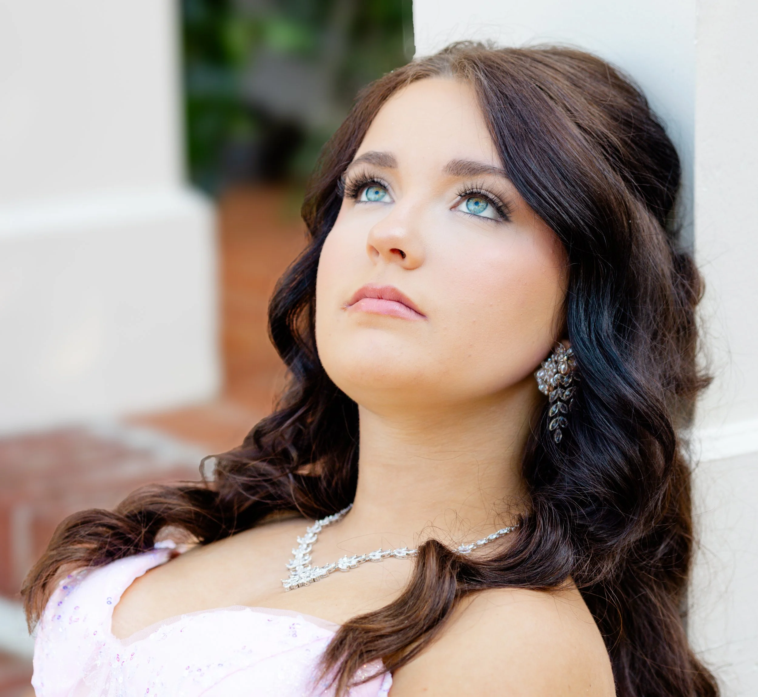 Close-up of a woman with blue eyes and long dark hair styled in loose waves, wearing bridal jewelry, standing near a white wall with greenery in the background.