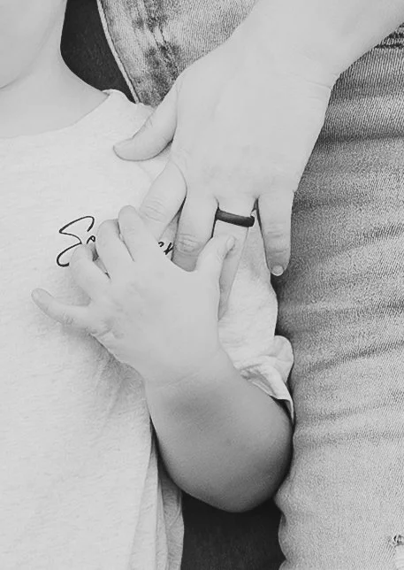 A person's hand gently holding a baby's hand, with the baby's thumb touching the person's finger, in black and white.
