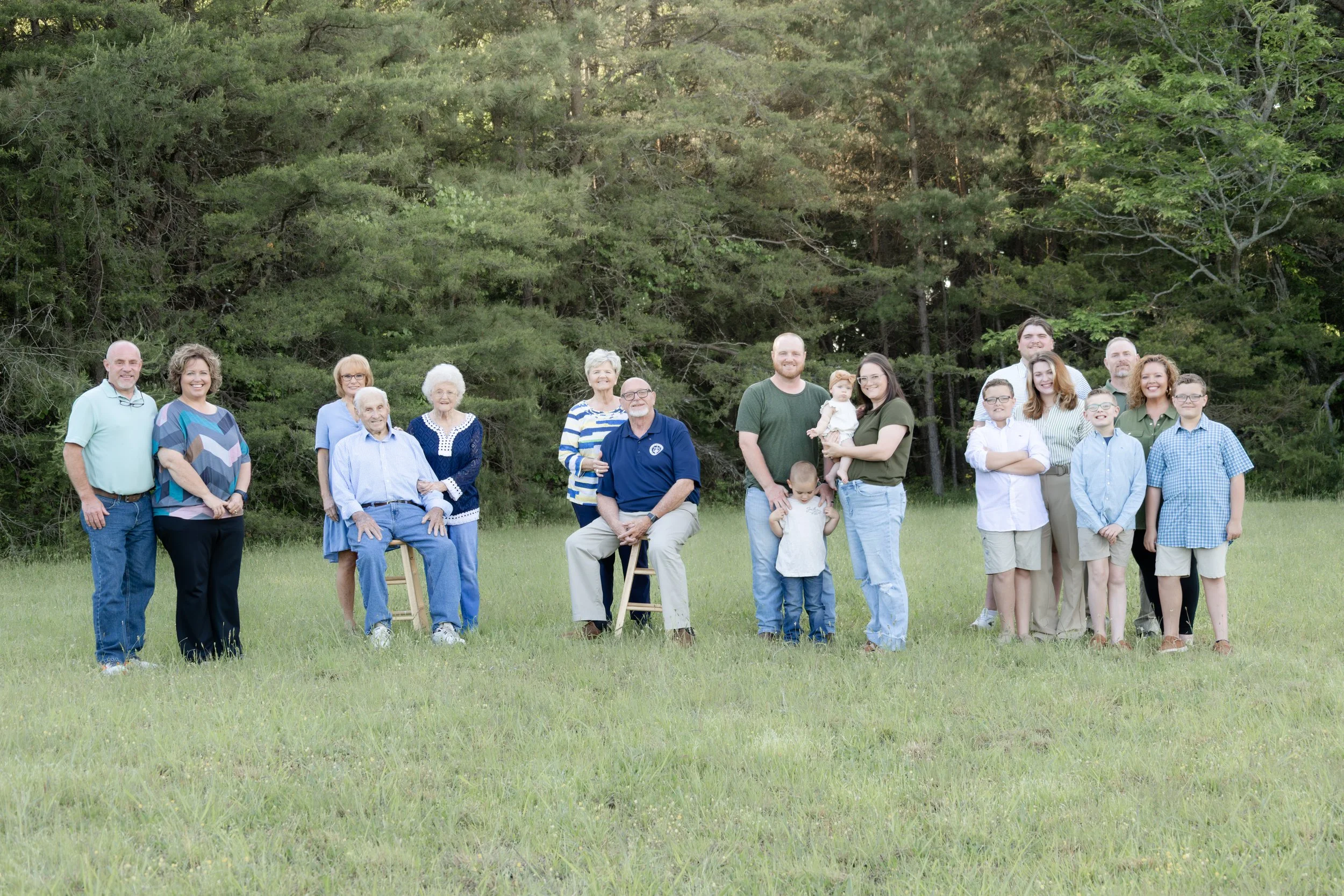 A large group of people, including elderly, adults, teenagers, and children, smiling outdoors in front of a wooded area with green trees and grass.
