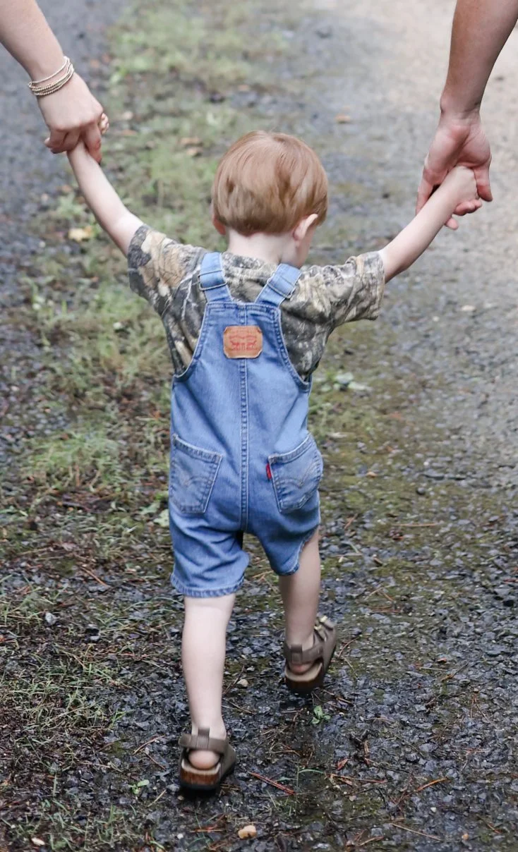A young boy with red hair wearing a camouflage shirt and denim overalls walking on a dirt trail, holding hands with two adults, one on each side.